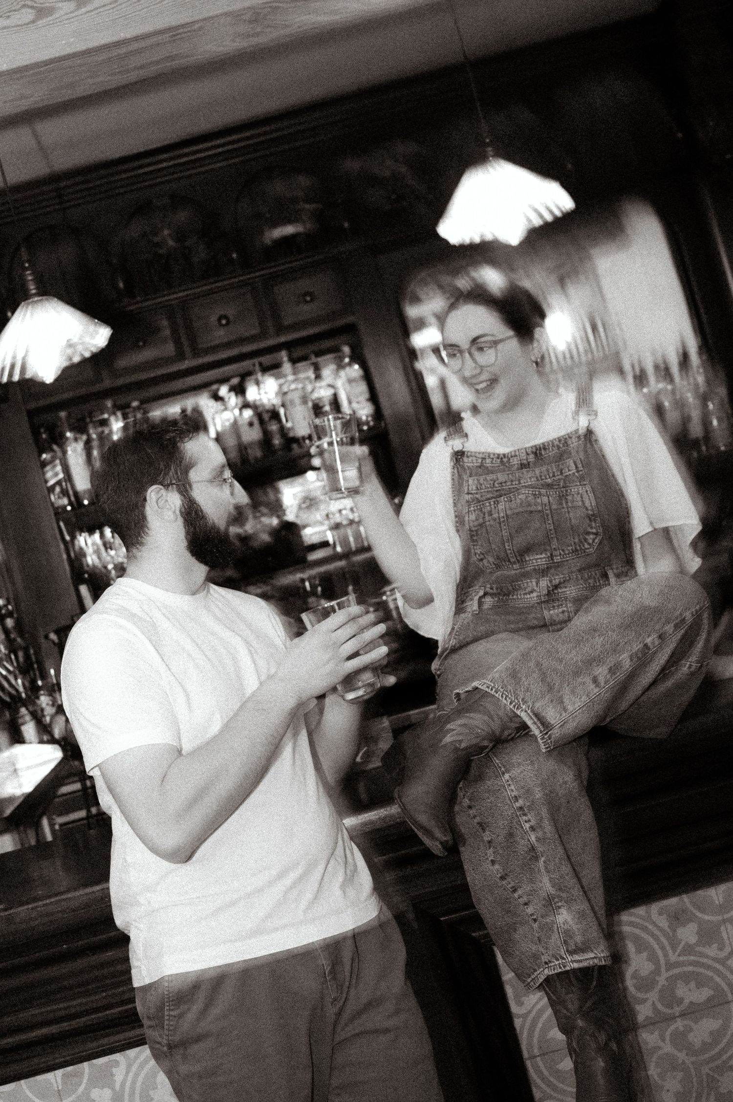 Couple laughing together at a bar, woman in denim overalls sitting on counter as man holds a drink.