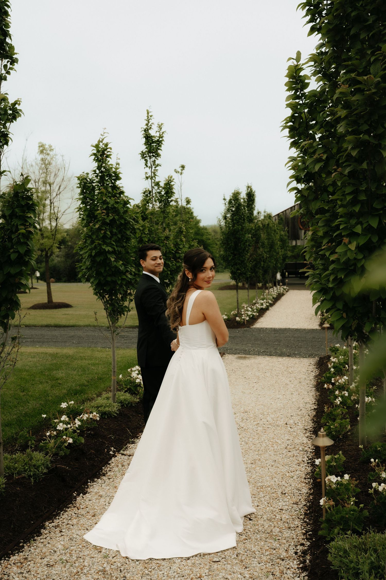 Bride and groom pose on a gravel garden path lined with tall hedges and white flowers at their outdoor wedding.
