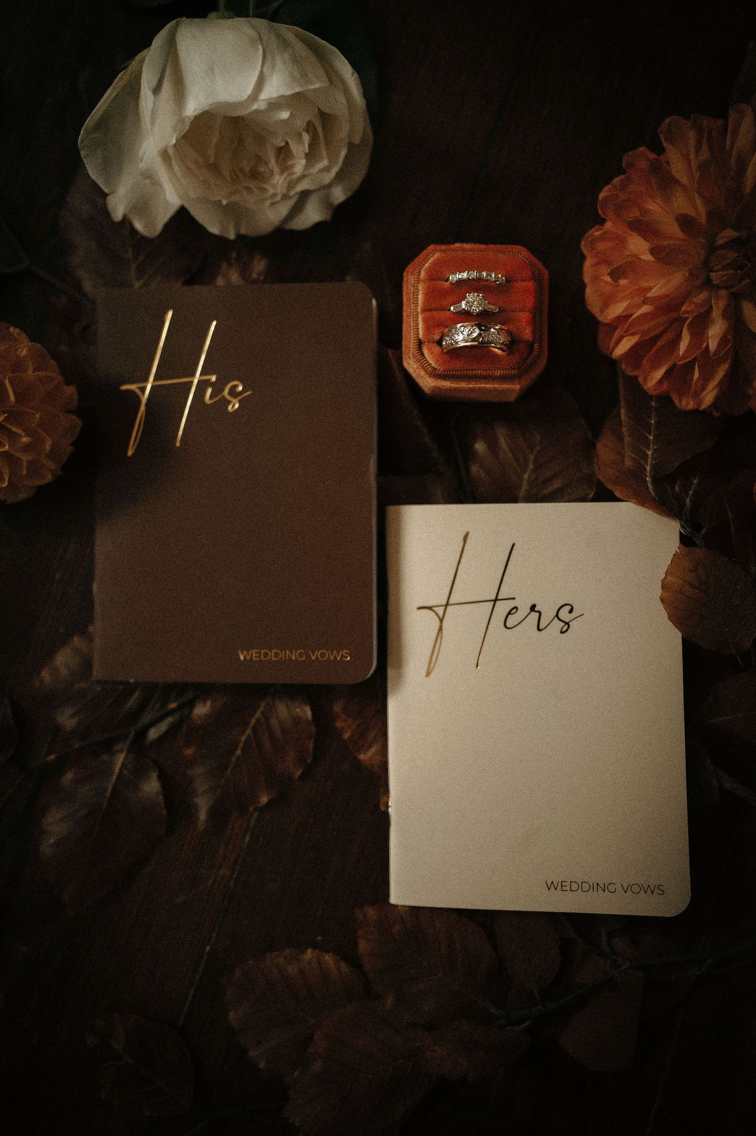Dark and moody wedding vow books with 'His & Hers' text surrounded by flowers.