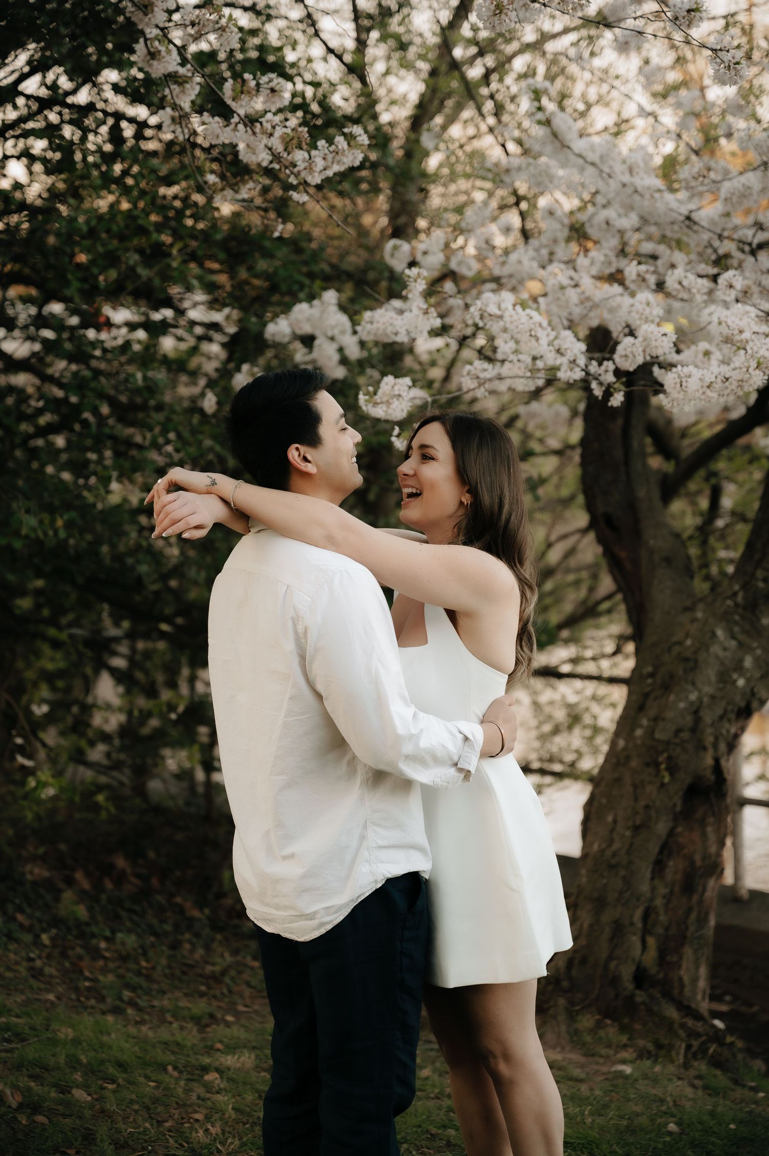 Couple embracing and laughing under blooming cherry blossom tree, wearing white outfits in a romantic outdoor setting.