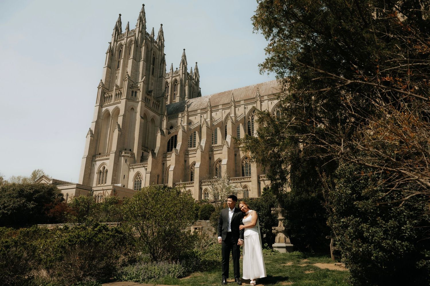 Couple poses in garden before grand Gothic cathedral on sunny day.