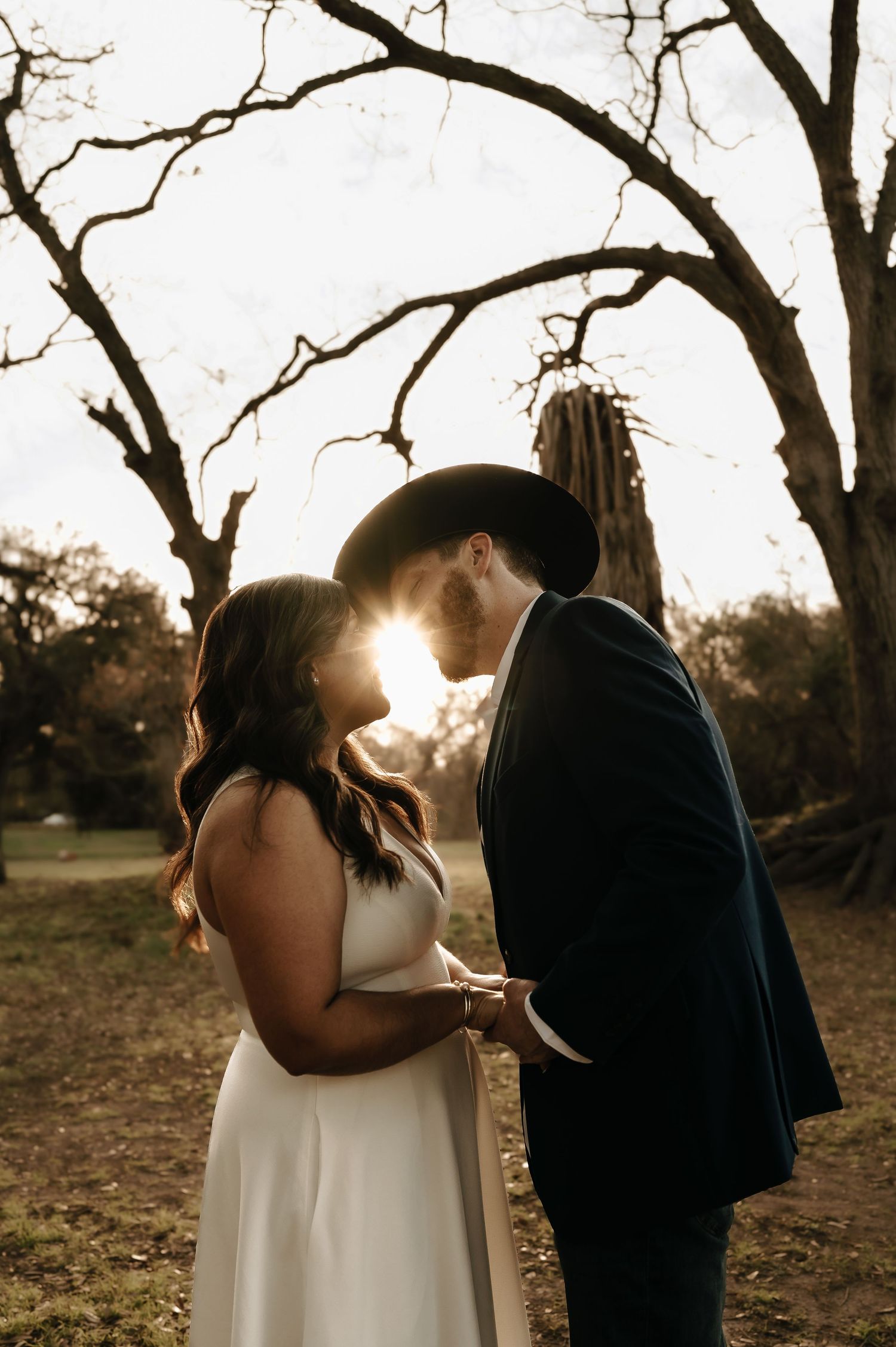 A backlit couple shares an intimate moment among bare winter trees at sunset.