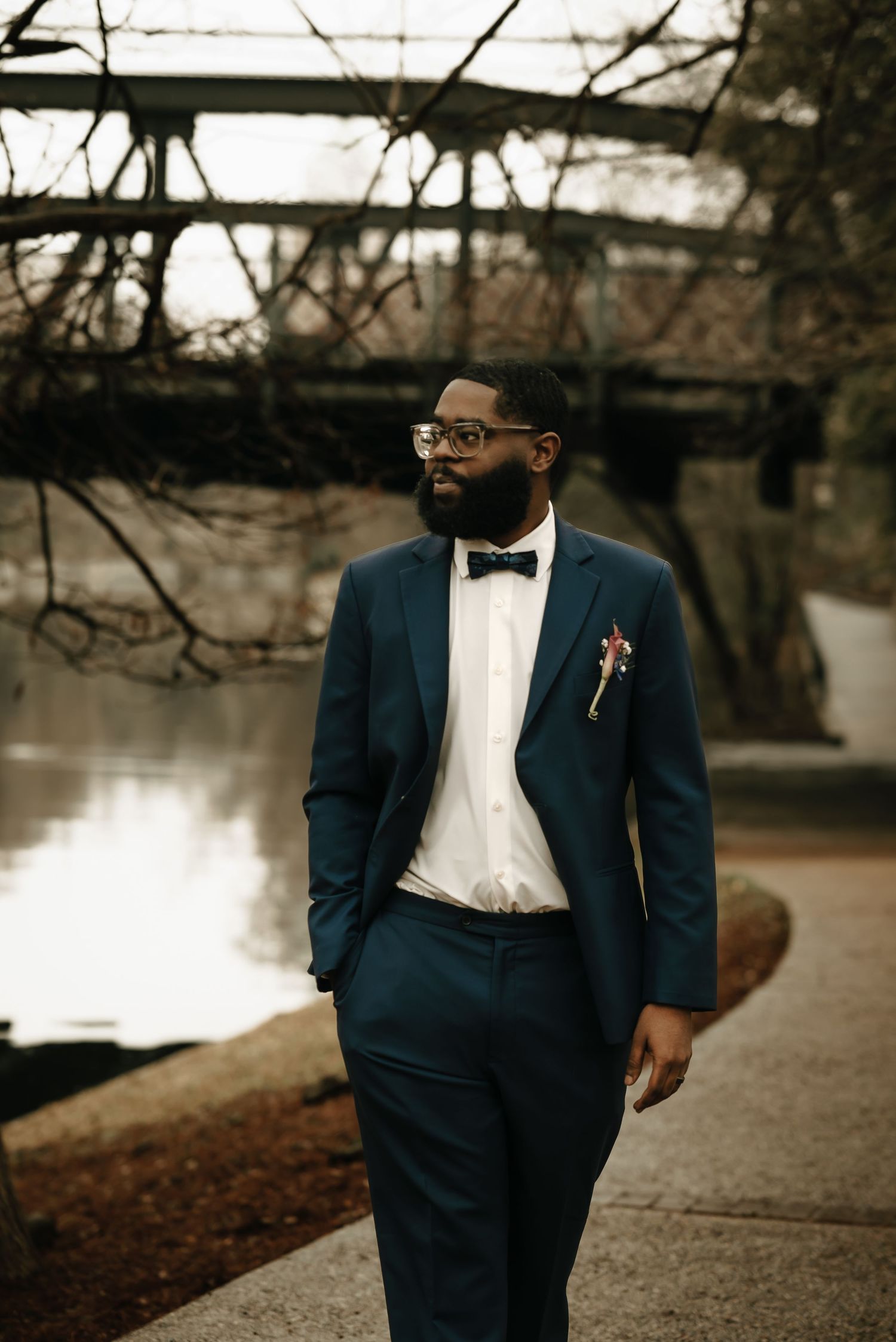 Groom in navy suit and bow tie poses thoughtfully by a lake during autumn.