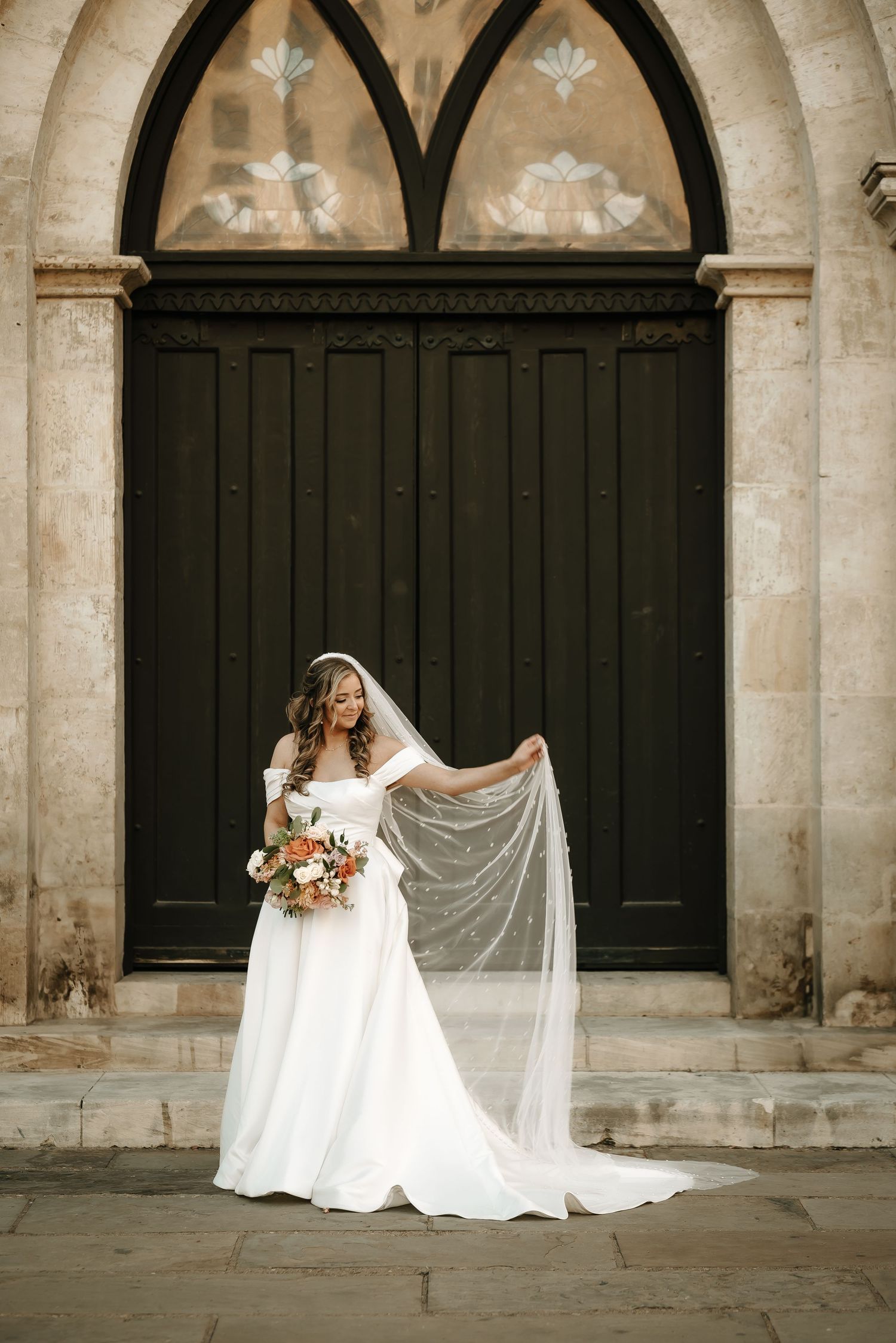 Bride with flowing veil poses in front of historic church architecture.