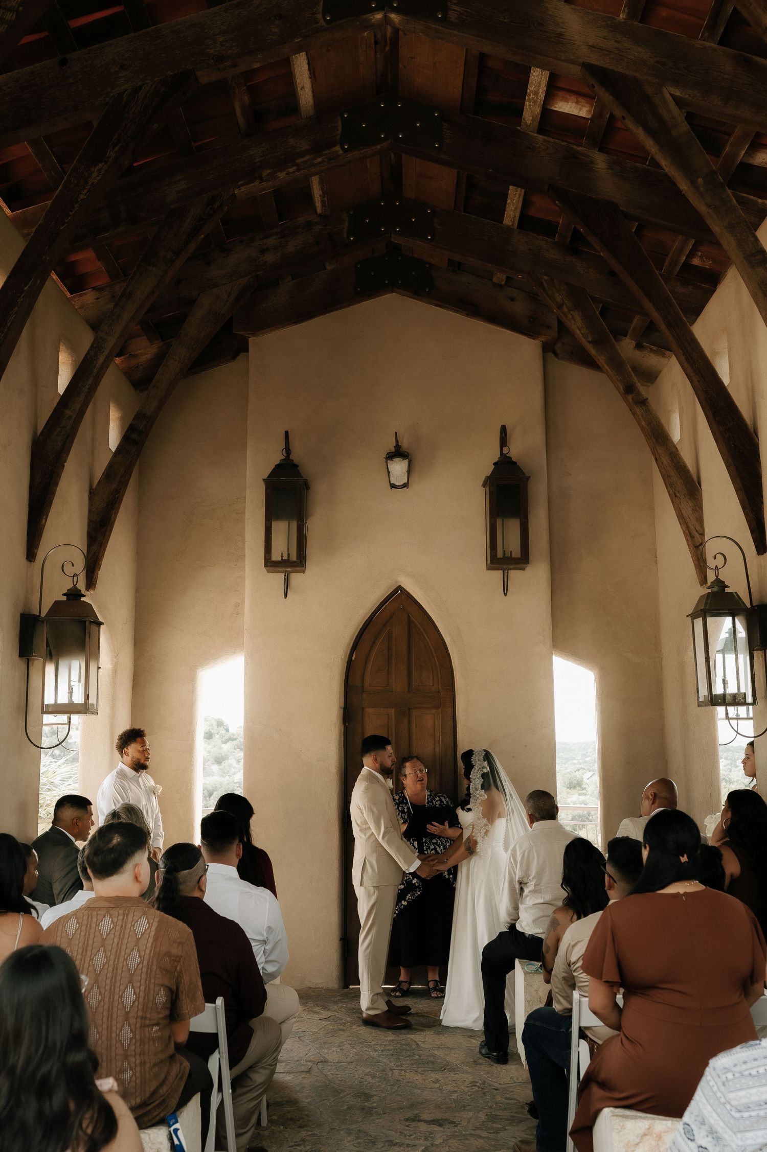 Wedding ceremony in progress inside a chapel with wooden beams and hanging lanterns.
