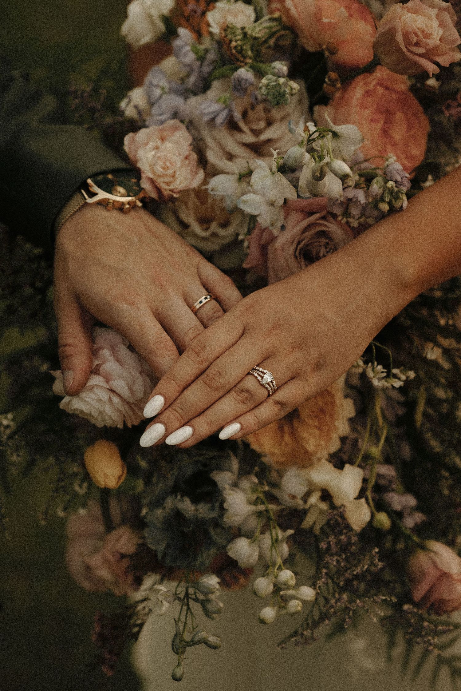Wedding rings displayed on hands against a peachy pink floral bouquet background.