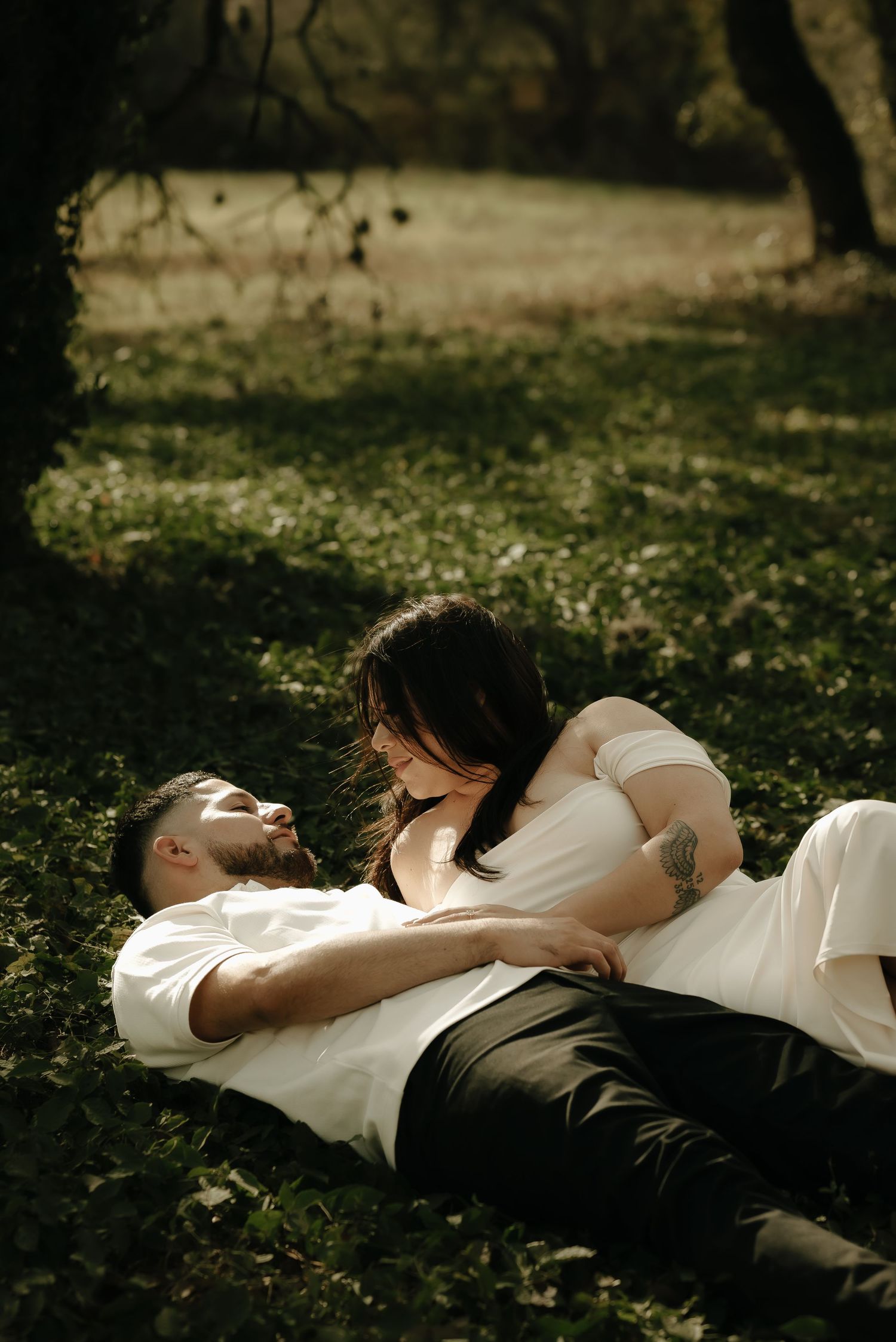 A couple in formal wear lies together on grass in dappled sunlight.
