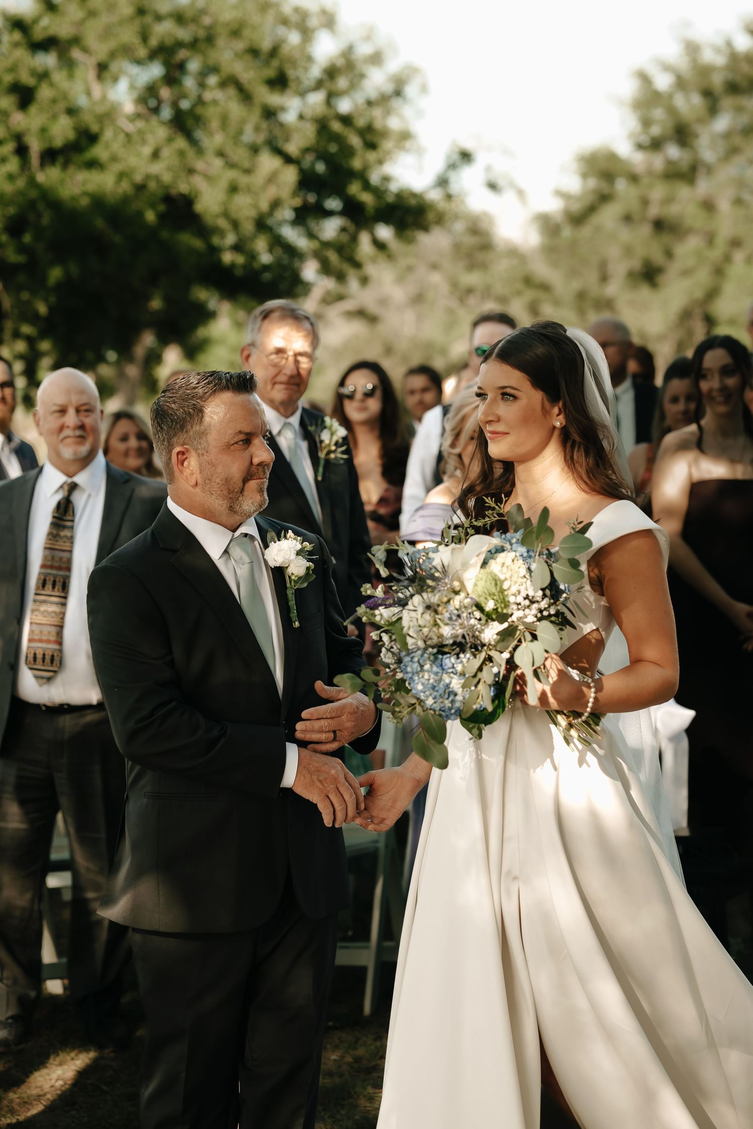 Wedding party gathered outdoors under trees with flowing white dresses and black suits creating a joyful atmosphere.