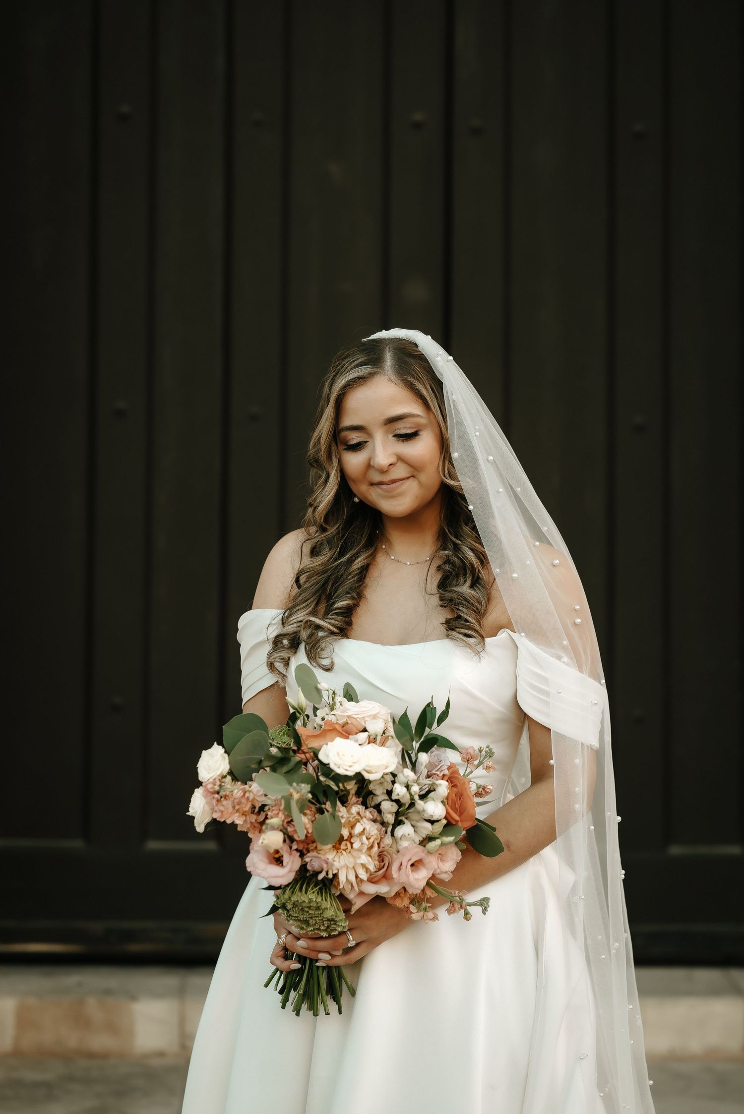 A bride in a white wedding dress holds a beautiful peach and pink floral bouquet against a dark background.