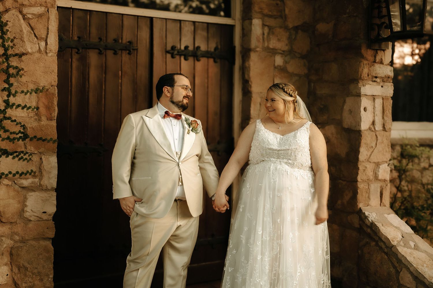 Elegant couple in wedding attire poses against a rustic stone wall backdrop.