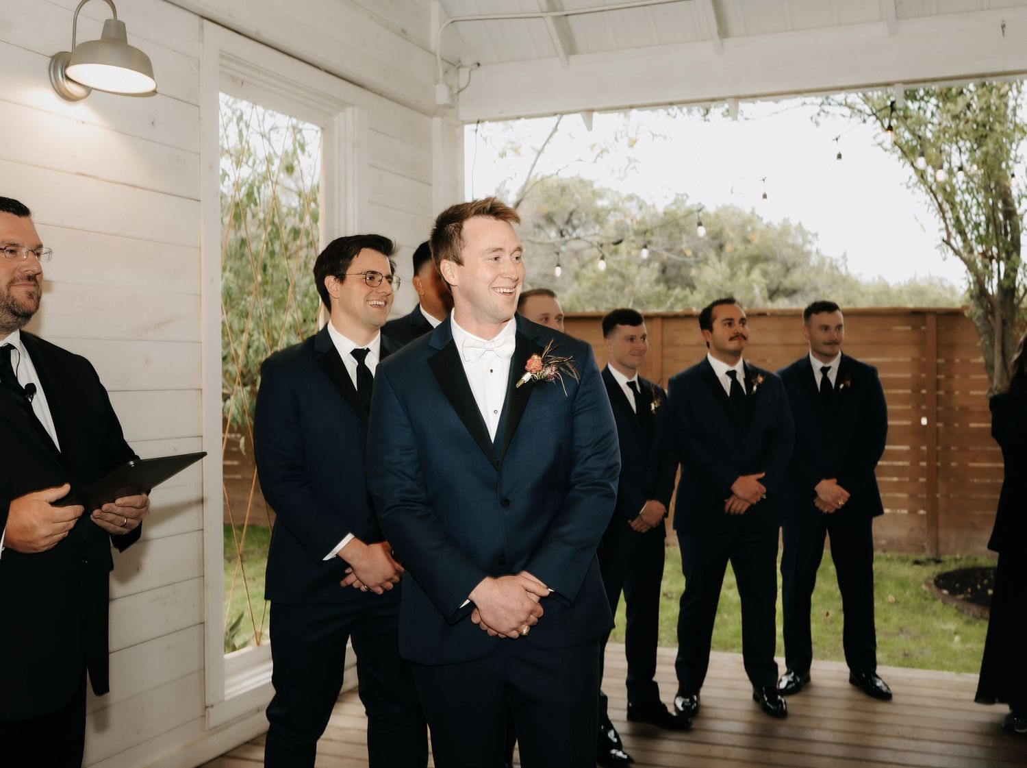 Wedding party in formal attire stands in line against white walls with elegant chandeliers overhead.