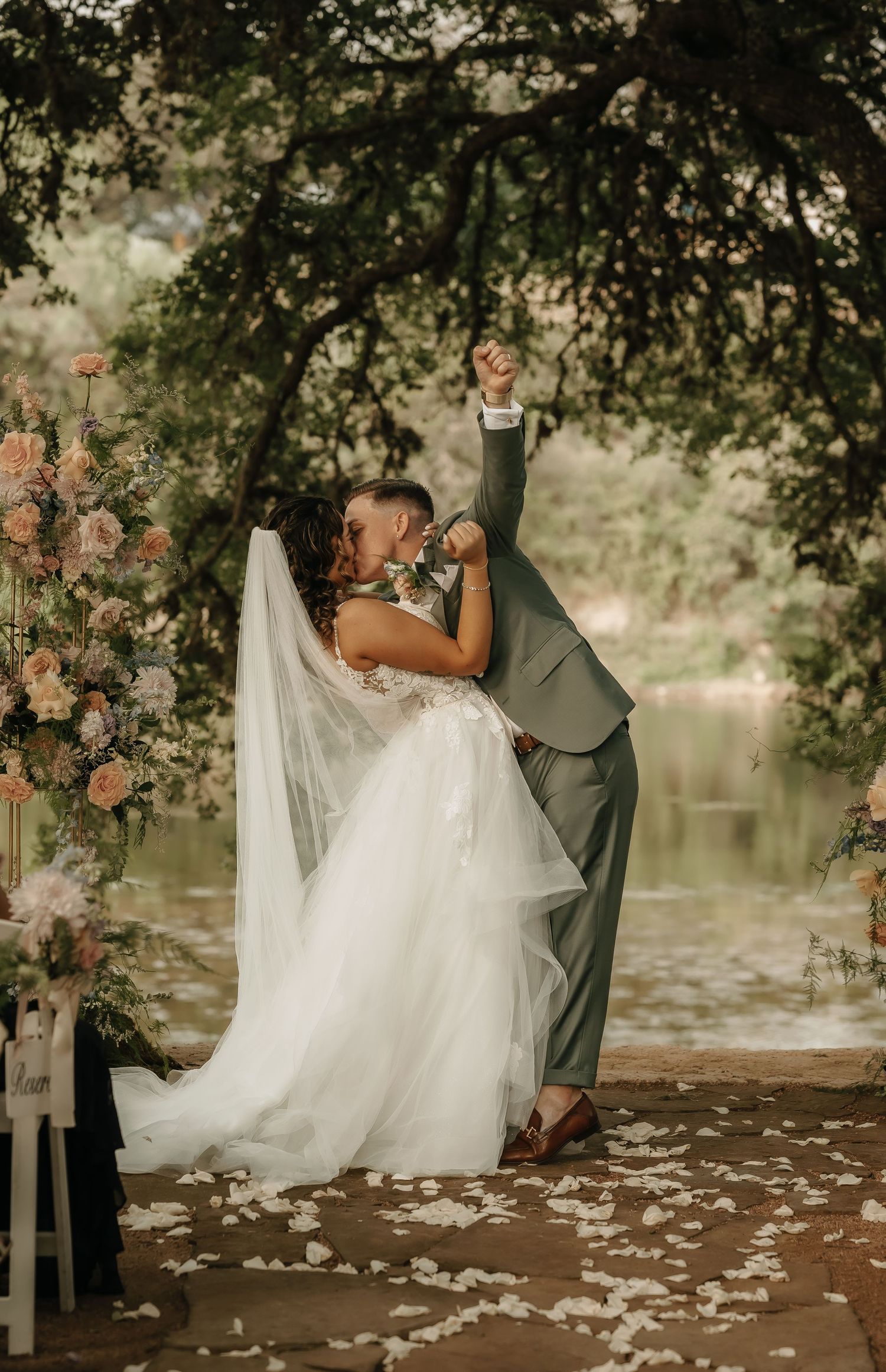 Romantic dip kiss under tree branches with floral arrangements and lake backdrop.