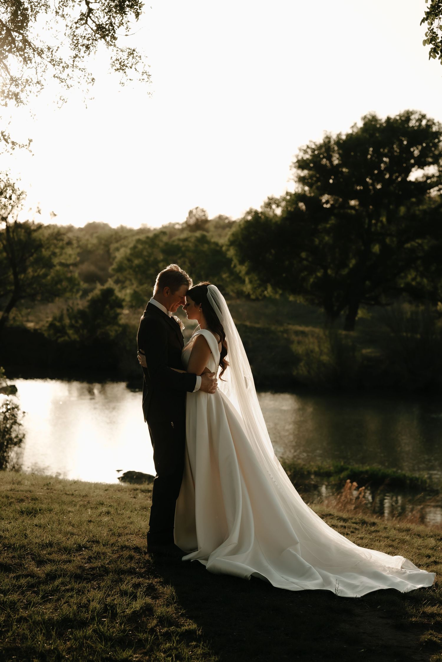 Newlyweds share a romantic moment by a lake at sunset with a flowing white wedding dress.