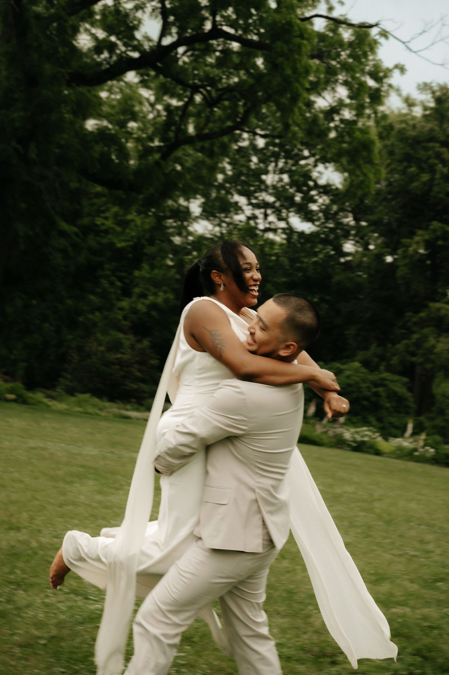 Joyful couple in white wedding attire embracing outdoors, groom lifting bride on lush green lawn.