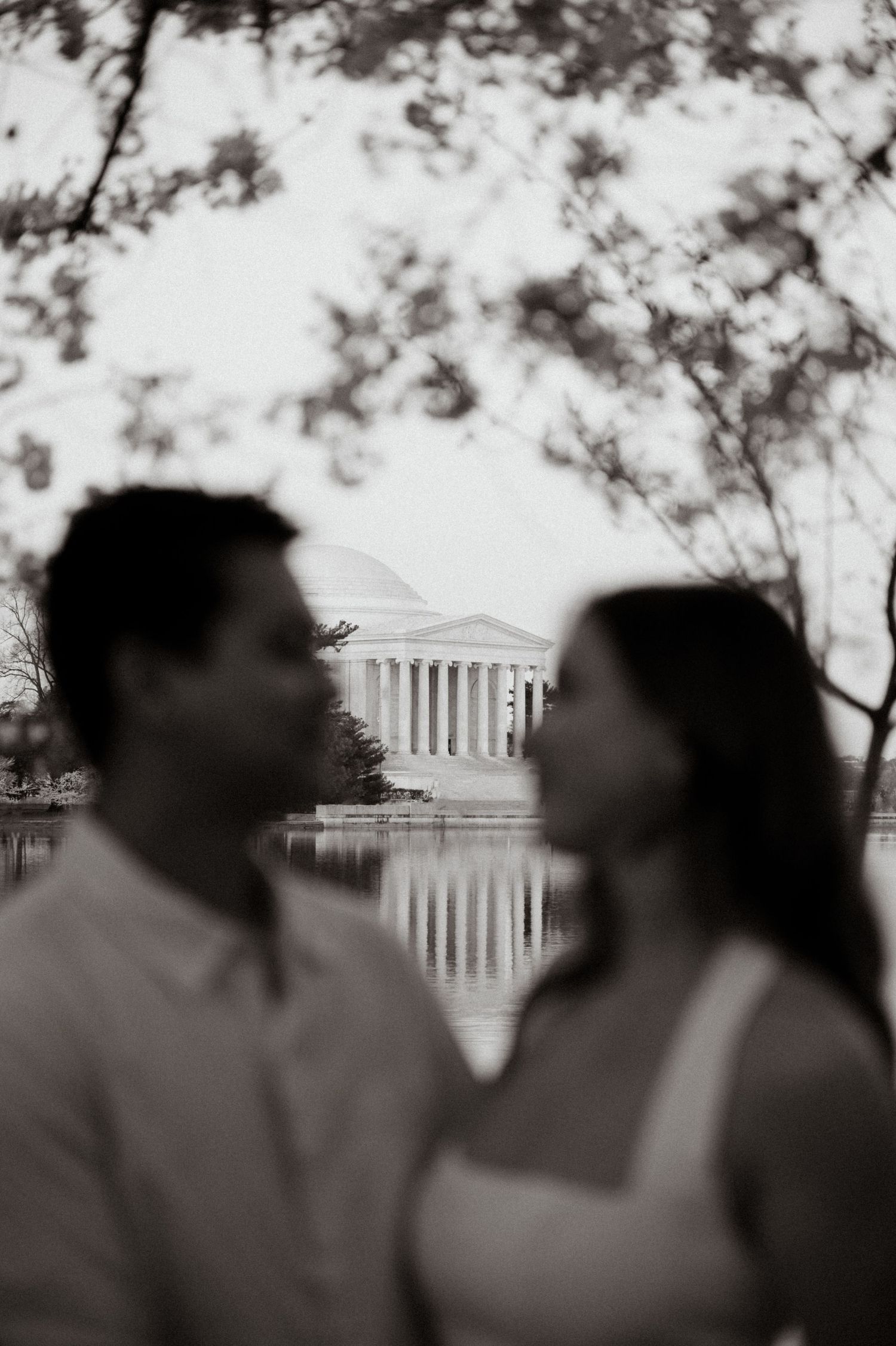 Black and white photo of a couple facing each other with the Jefferson Memorial reflected in water behind them.