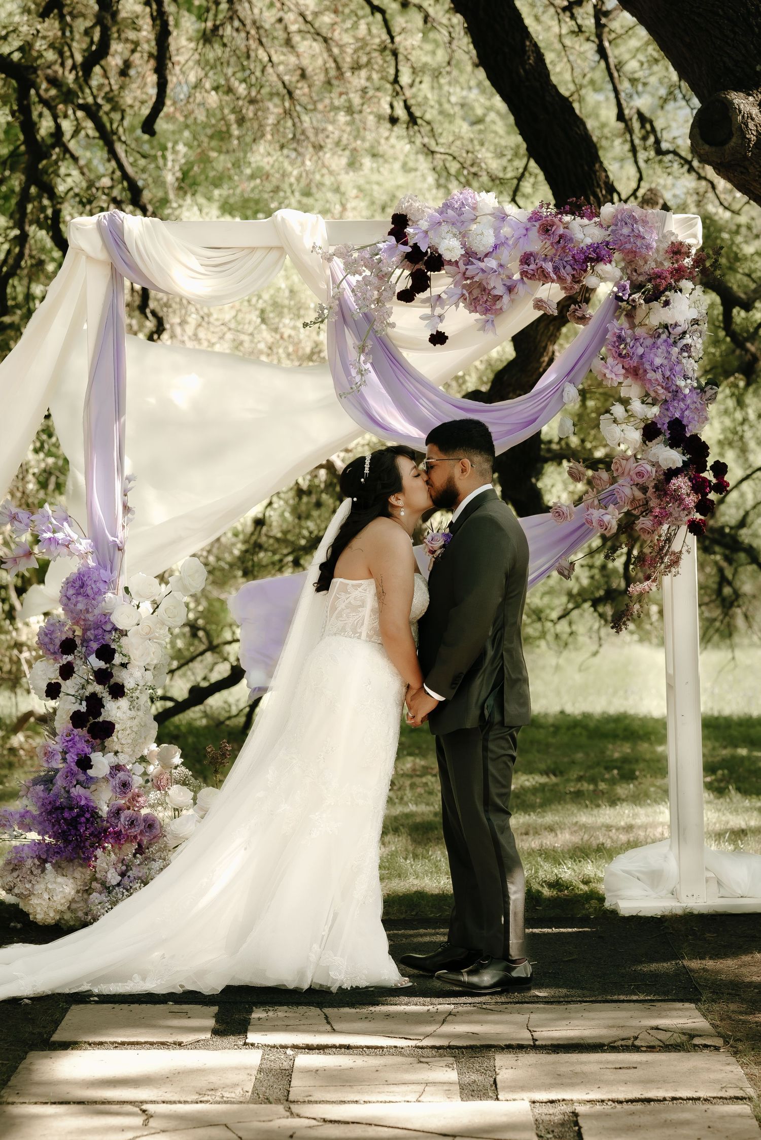 Outdoor wedding ceremony under elegant white and purple floral arch with flowing fabric.
