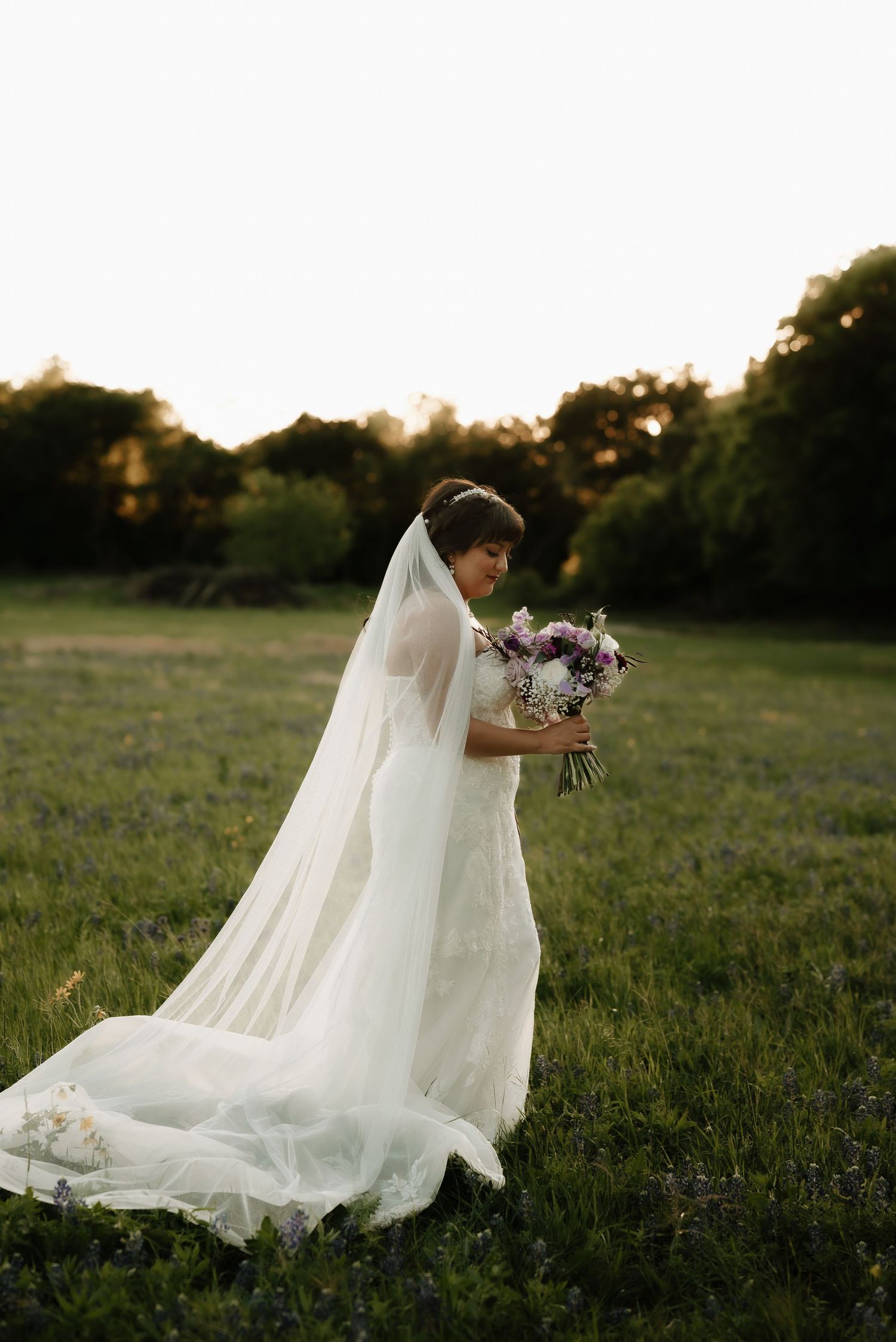 Flowing white wedding dress and veil trail across green field at sunset.