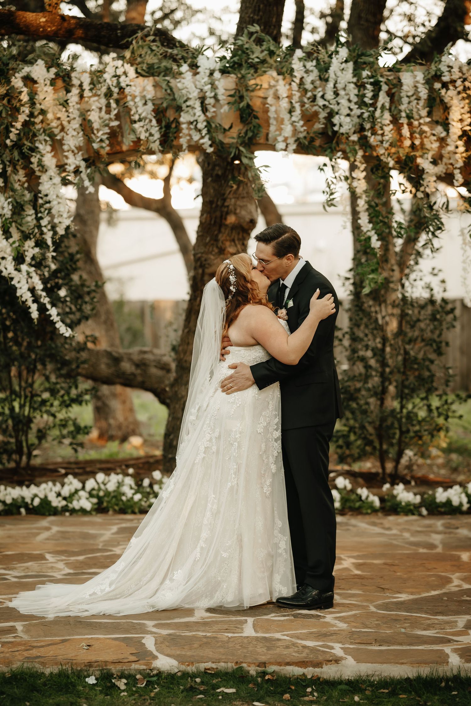 Newlyweds share intimate kiss under hanging floral decorations during outdoor wedding ceremony.