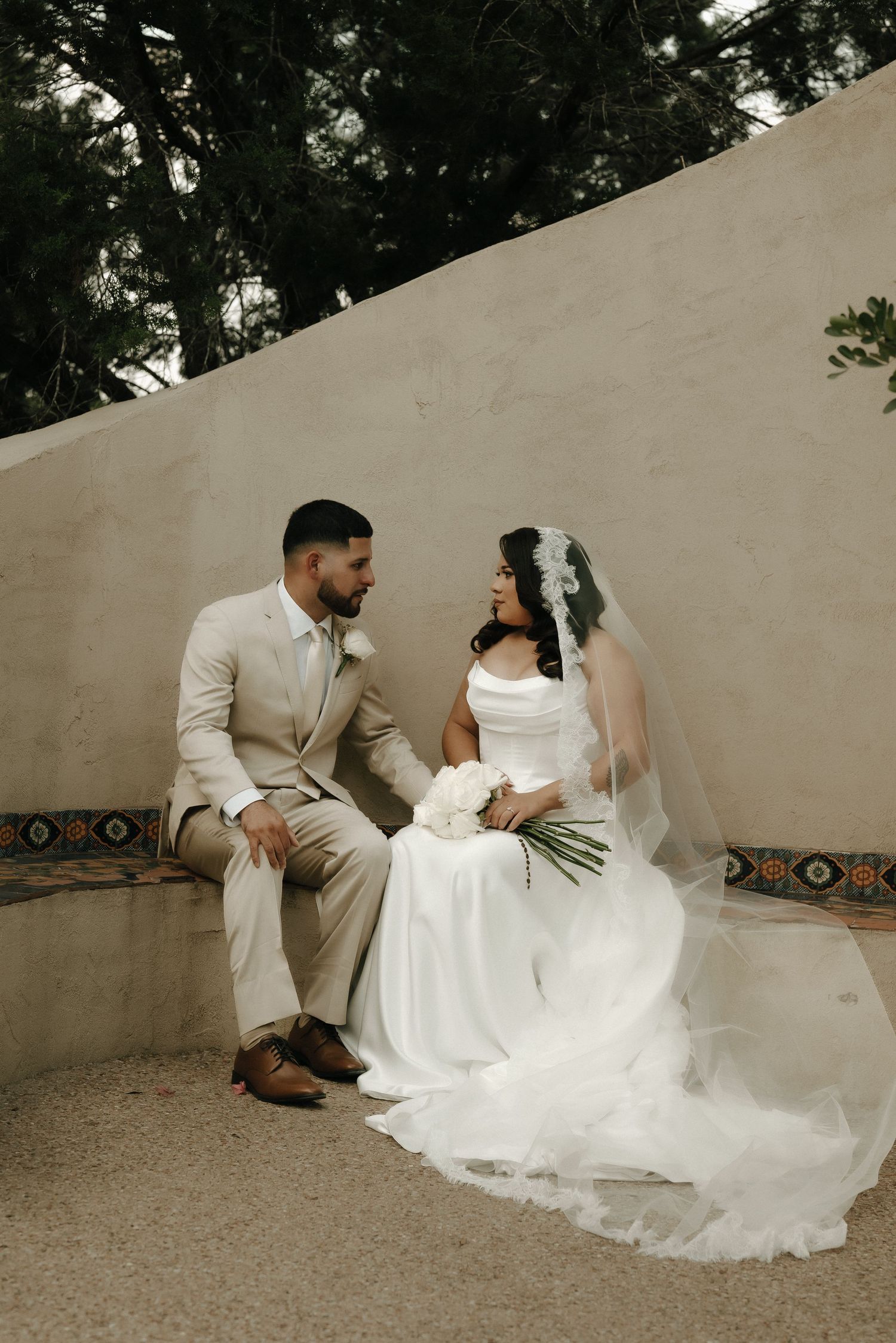 A couple in wedding attire sits on a decorative bench against a stucco wall.