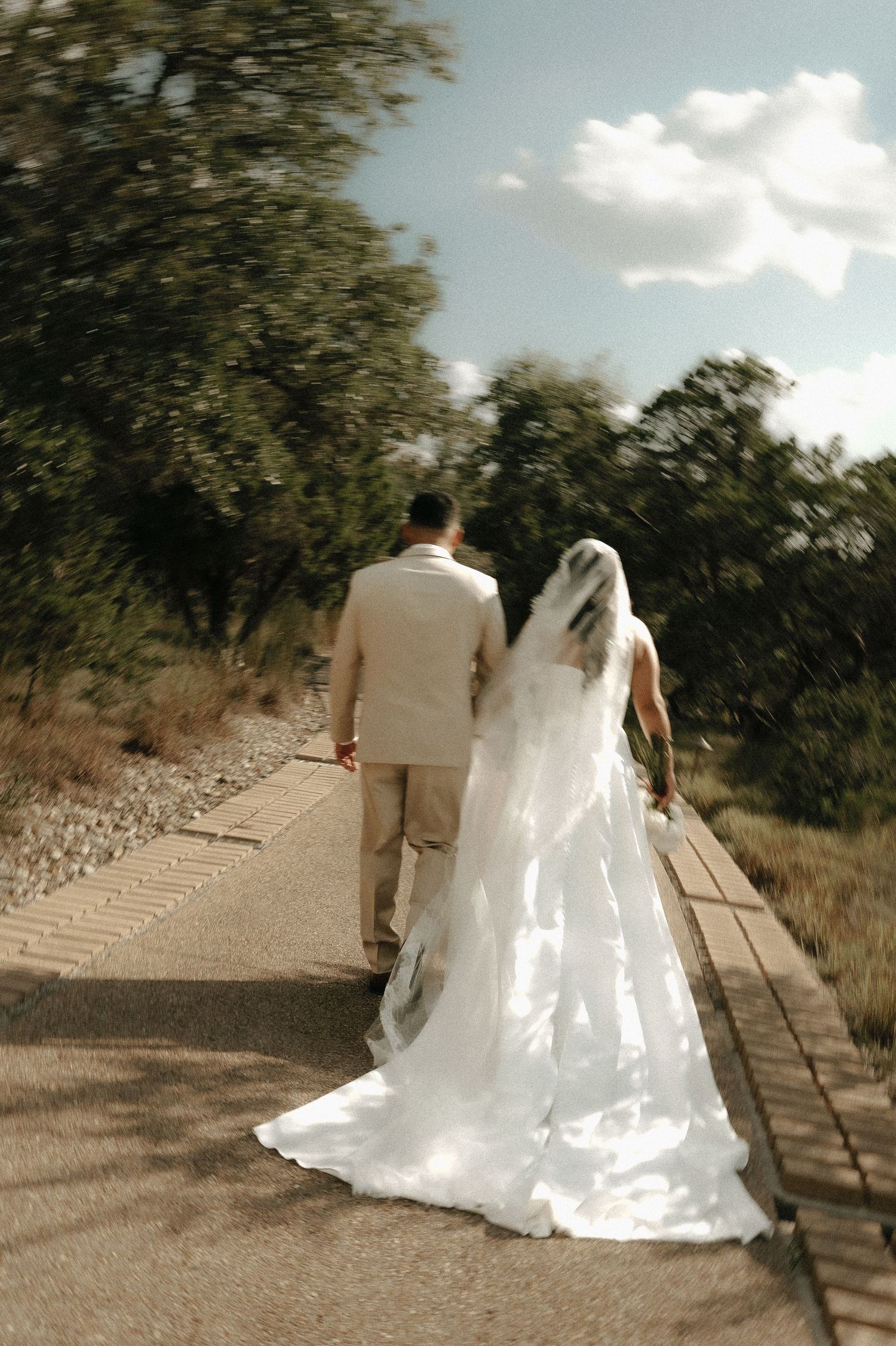 Bride and groom walking along a sunlit pathway surrounded by trees.