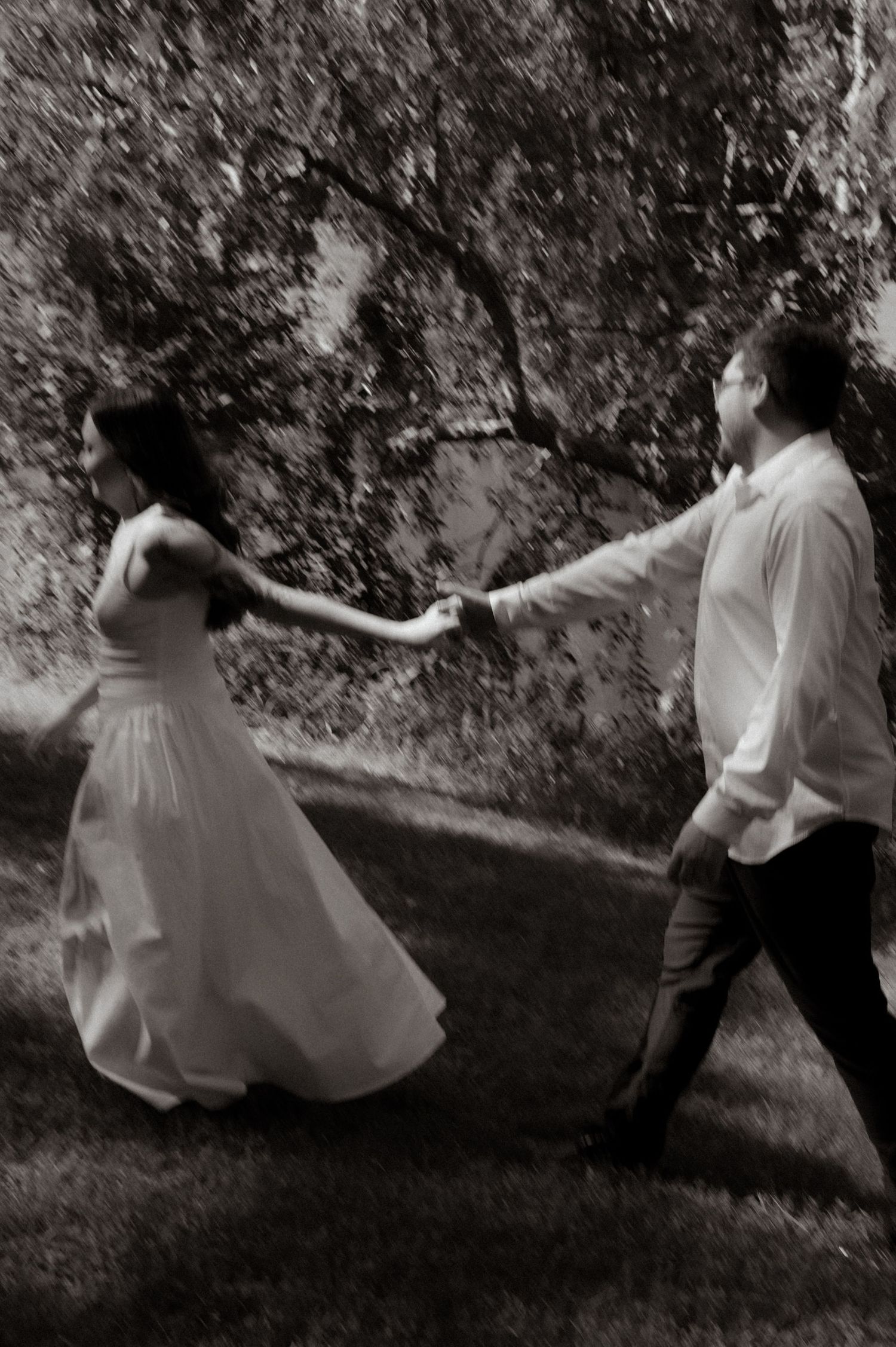 Black and white photo of a bride in white gown and groom holding hands, walking through a lush garden.