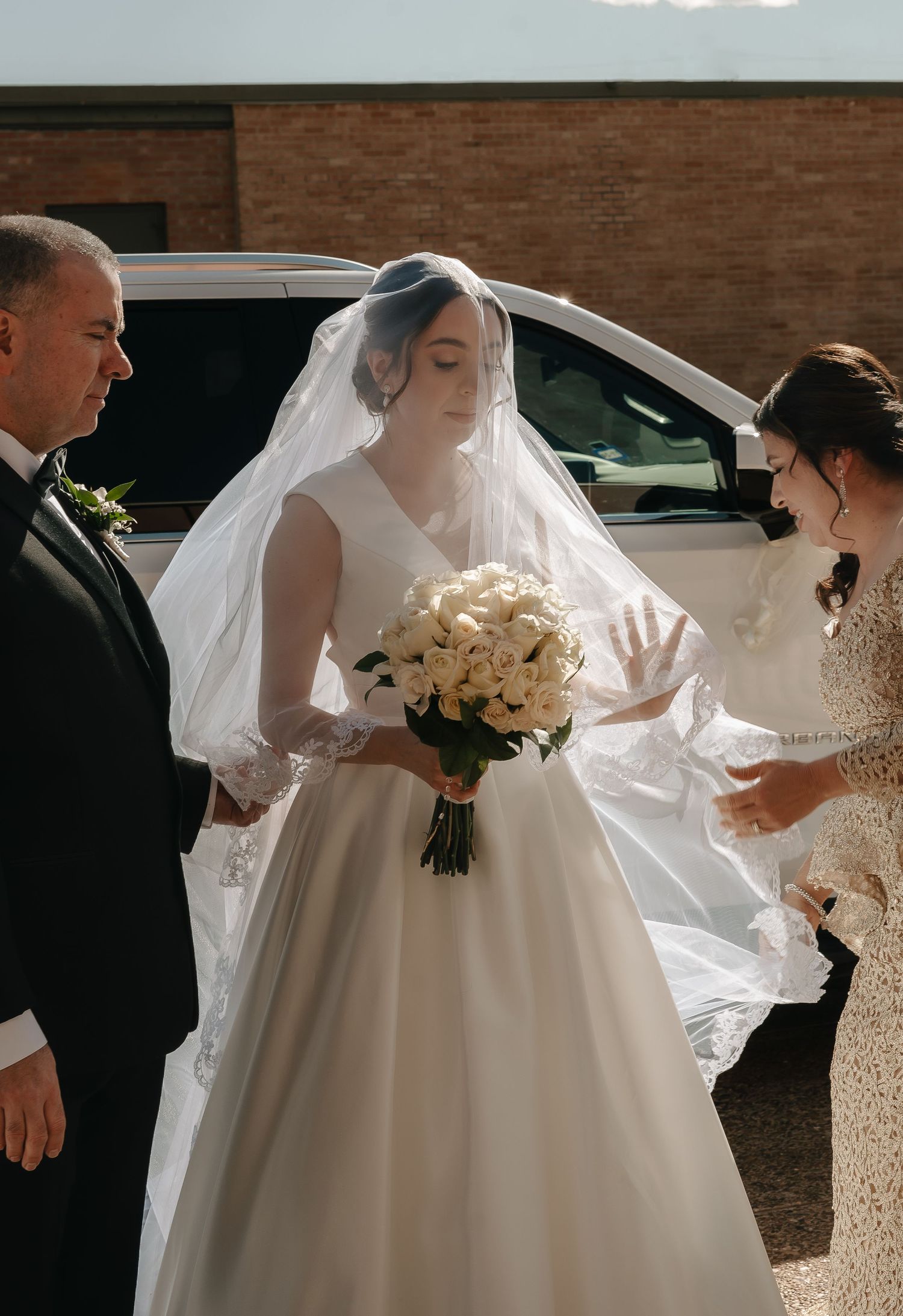 Elegant bride in a classic white gown holds a white bouquet while her veil flows in the breeze outside a brick building.