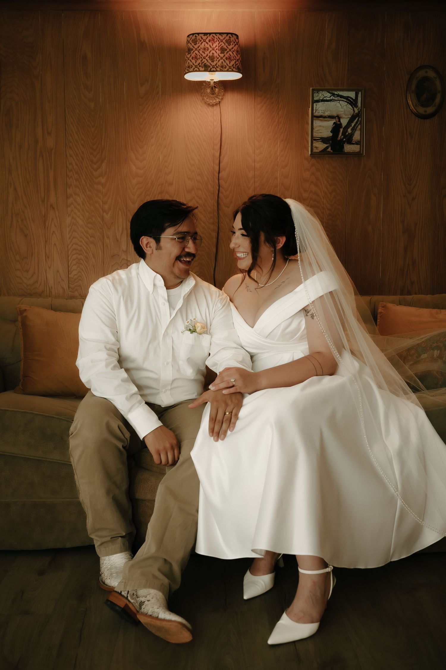 A couple in wedding attire sits intimately together on a wooden bench in a warm-toned indoor setting.