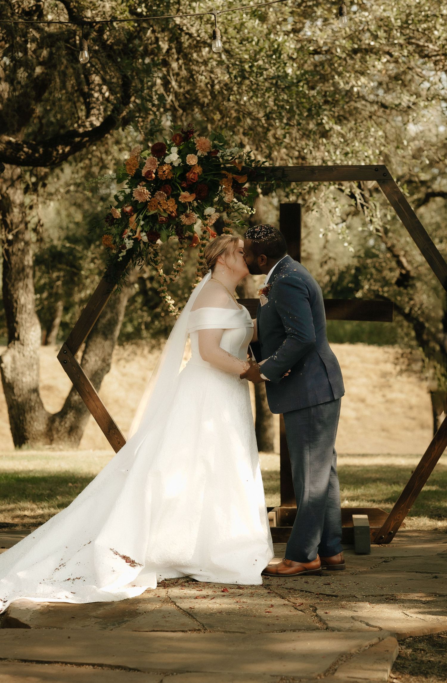 A wedding ceremony unfolds on a wooden platform under trees in warm sunlight.