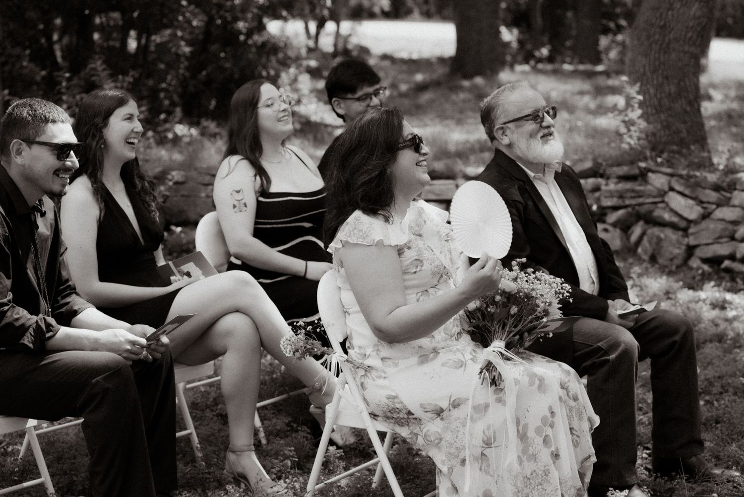 Wedding guests seated on white chairs watch the ceremony in a wooded outdoor setting.