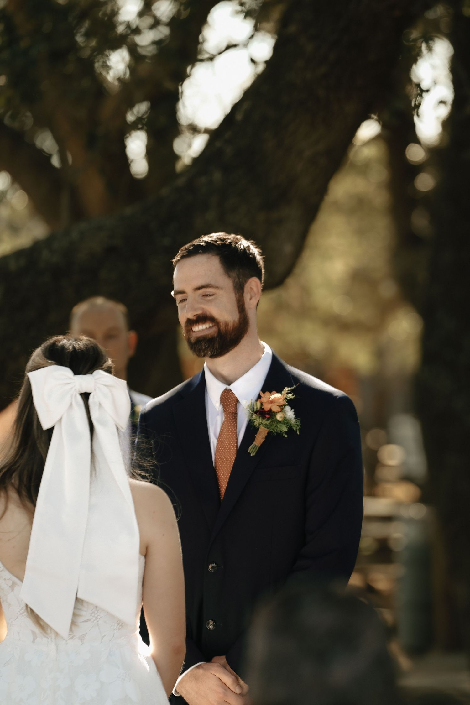An intimate wedding moment captured between a couple in formal attire under shaded trees.