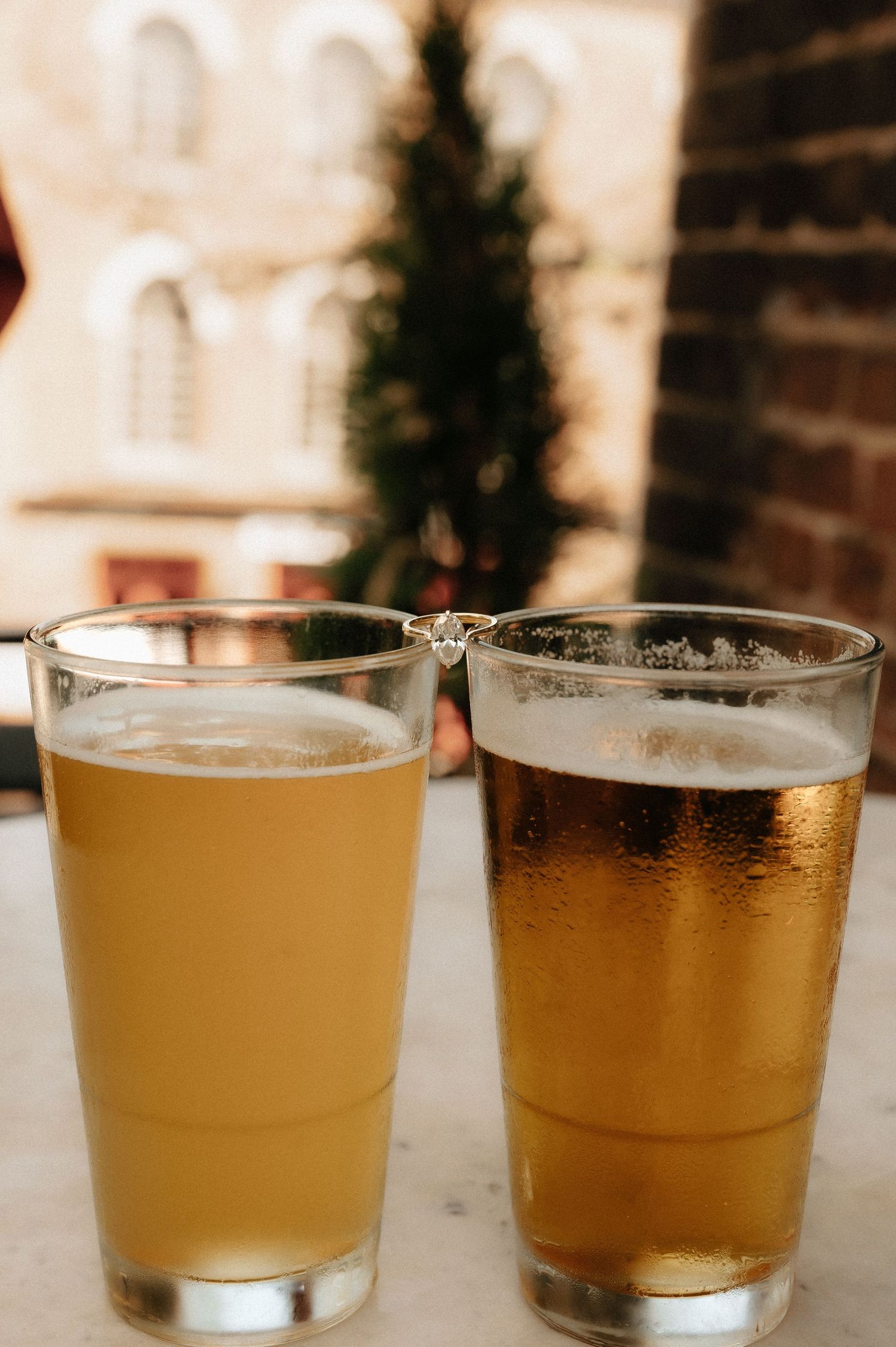 Two pint glasses of craft beer on a marble table with a blurred brick building and evergreen tree in the background.
