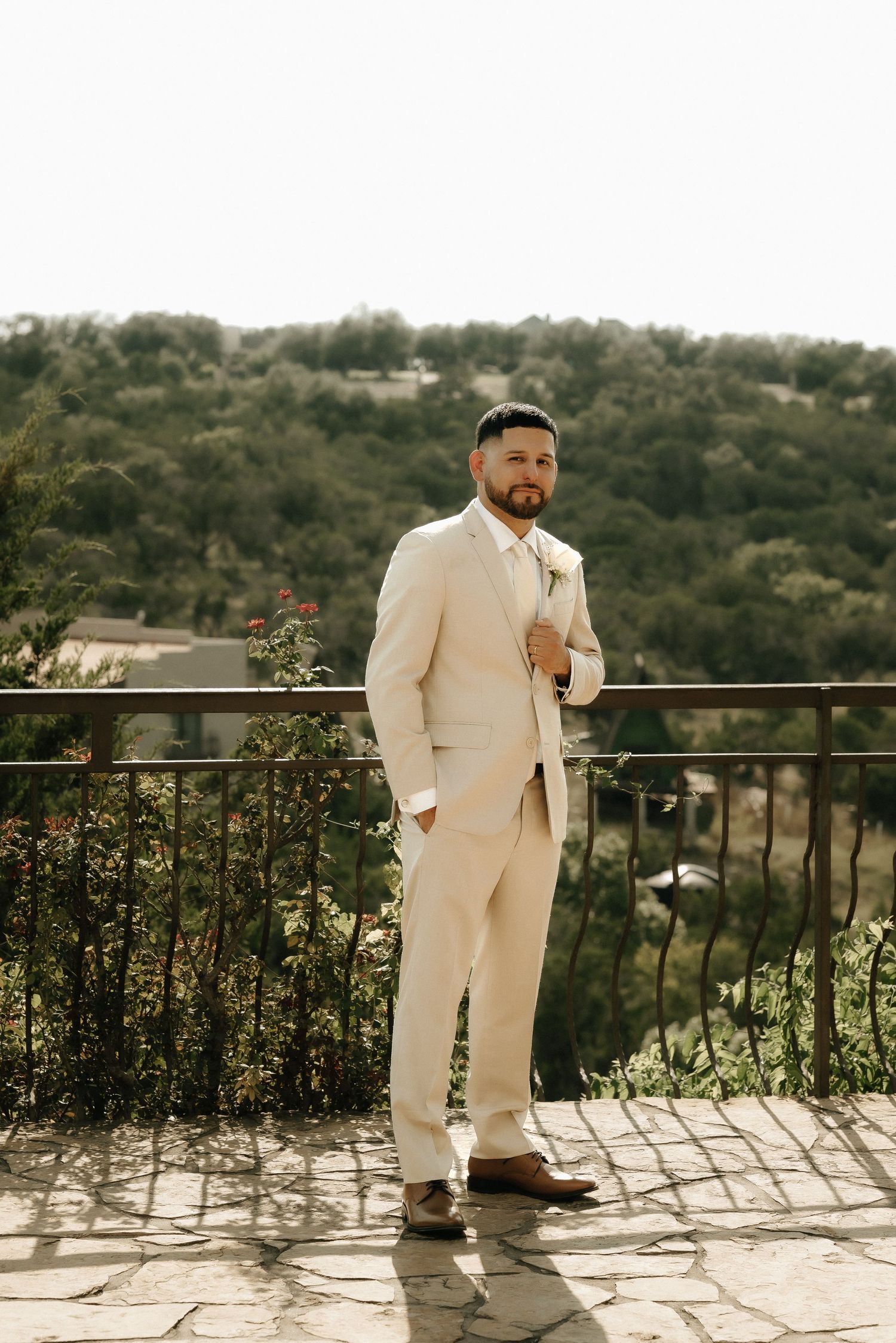 A groom in a cream-colored suit poses on a stone terrace overlooking a scenic hillside.