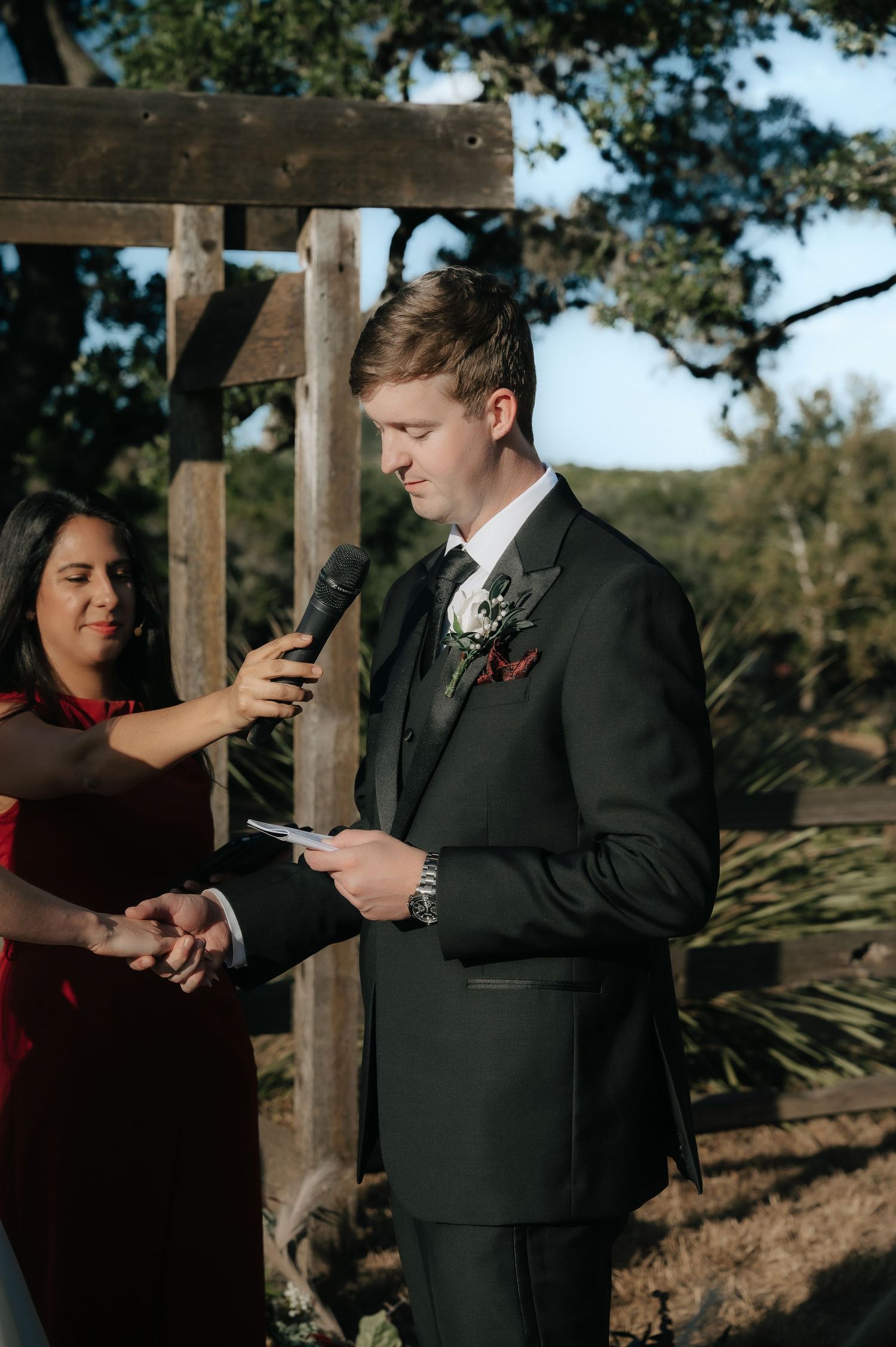 Groom in black suit reads from phone during outdoor wedding ceremony.