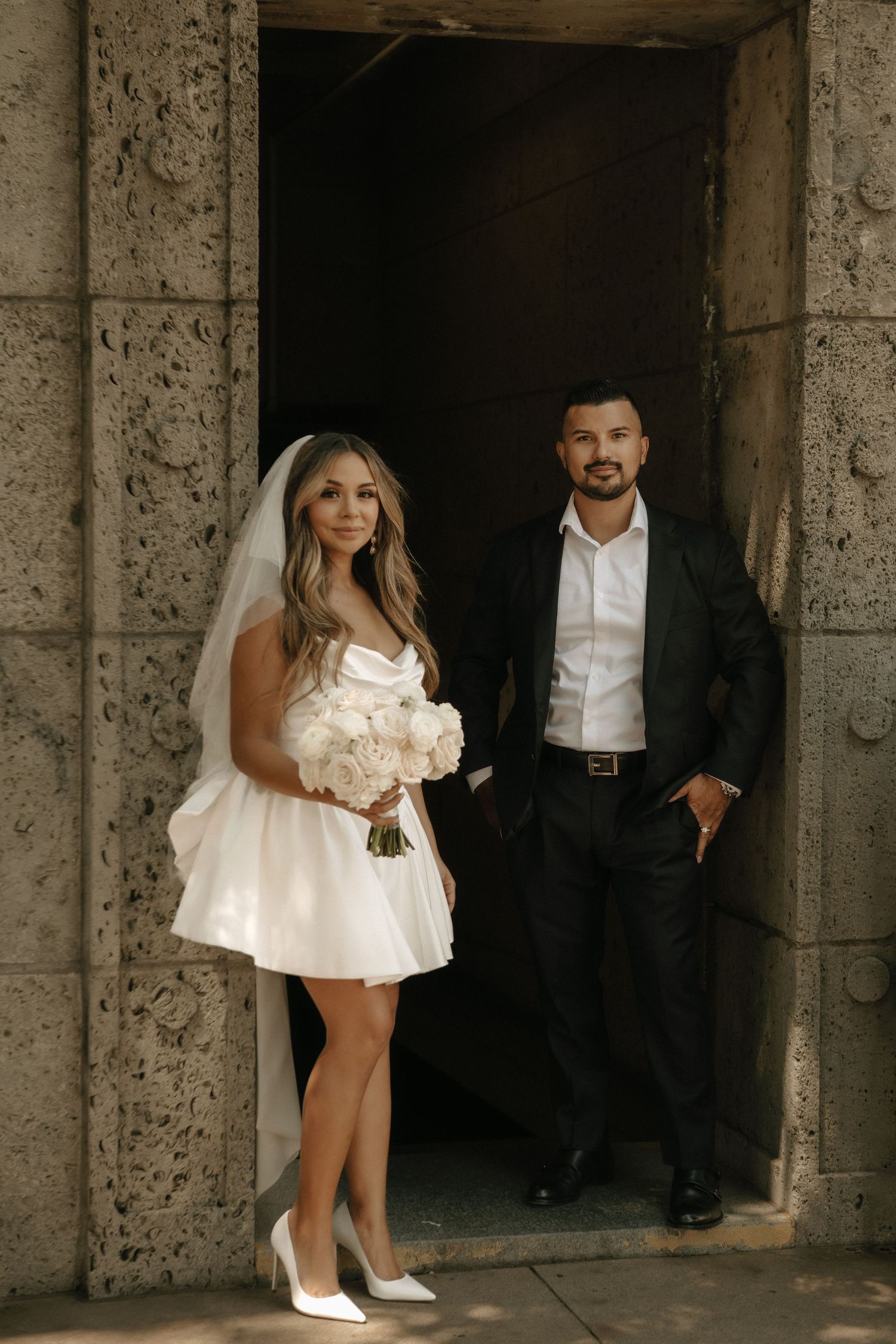 A couple poses in a stone doorway, with the bride wearing a short white dress and holding a bouquet.