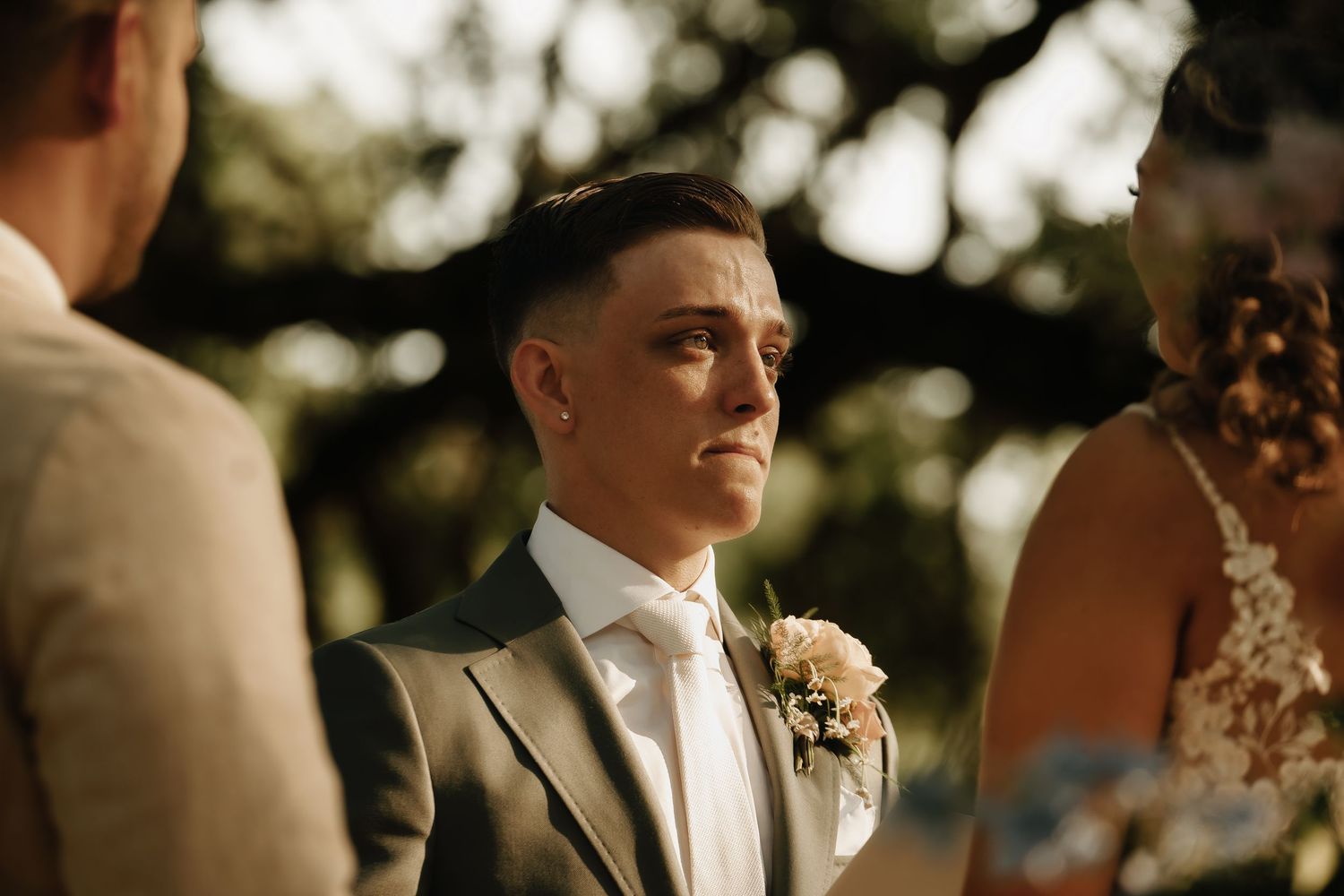 Wedding party members stand in formal attire during an outdoor ceremony under natural lighting.
