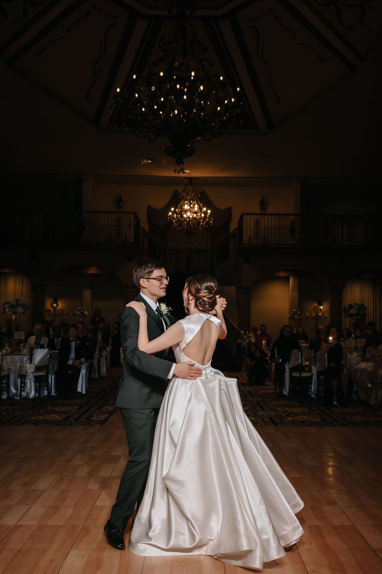 Couple sharing their first dance in an elegant ballroom with chandelier lighting.