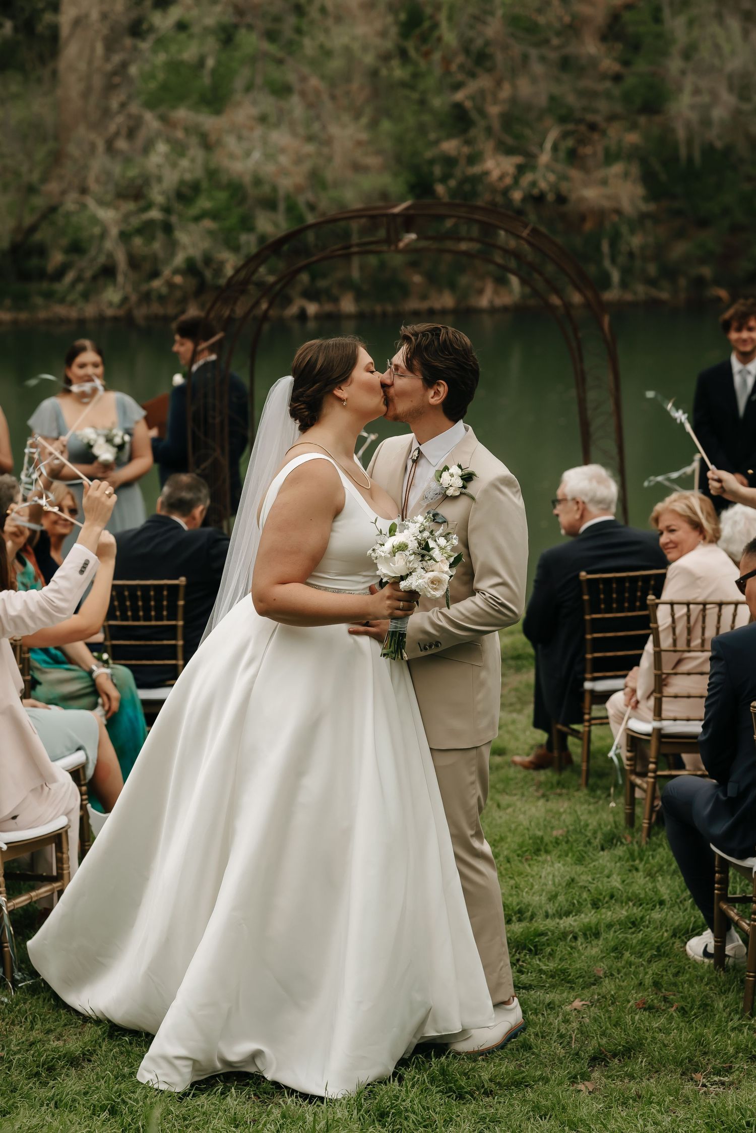 Romantic kiss during outdoor lakeside wedding ceremony with guests seated on lawn.