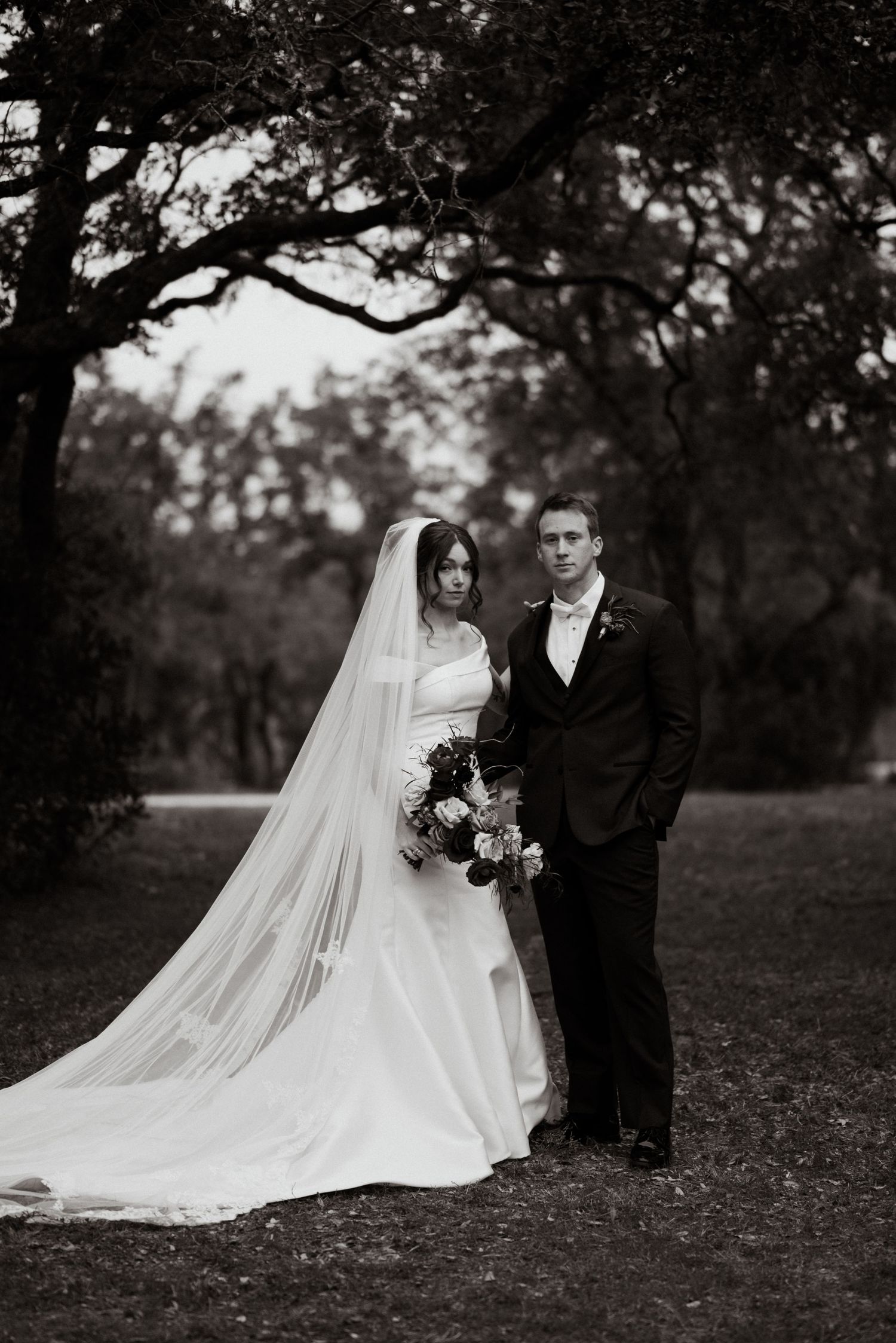 A black and white wedding portrait showing a bride's flowing veil against a natural outdoor backdrop.
