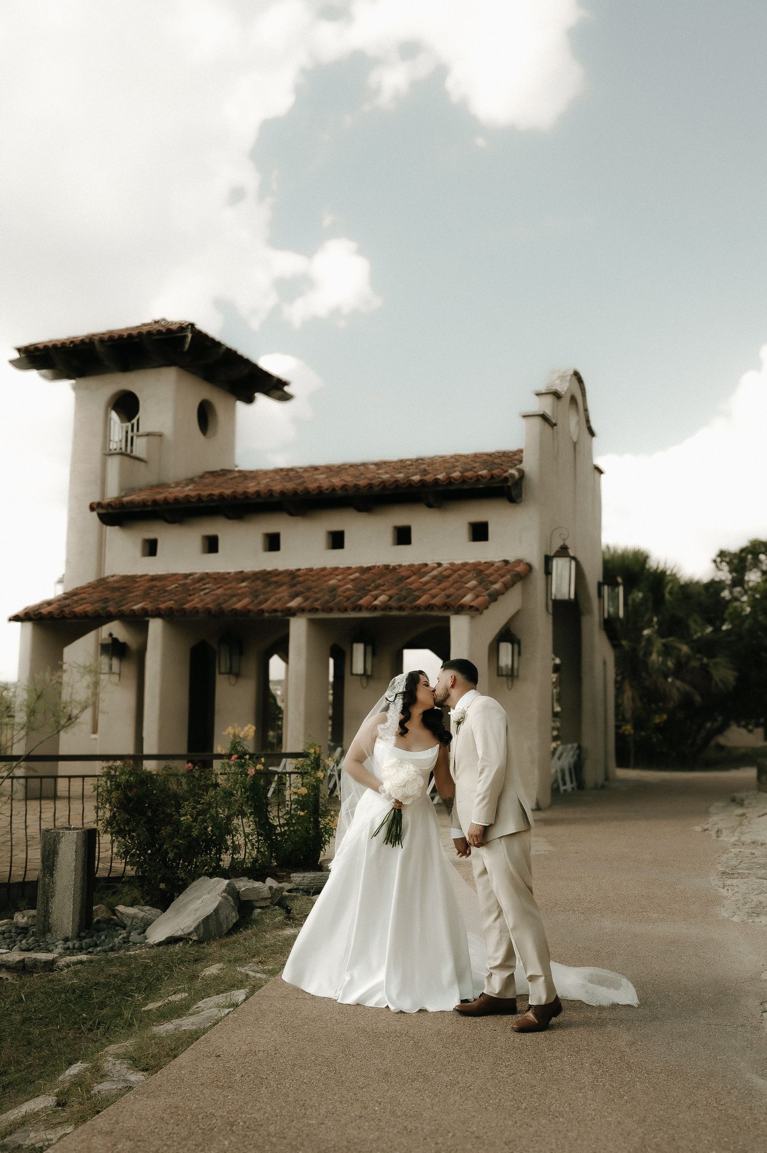 A wedding couple poses in front of a Spanish-style mission building with white stucco walls and terracotta roof tiles.