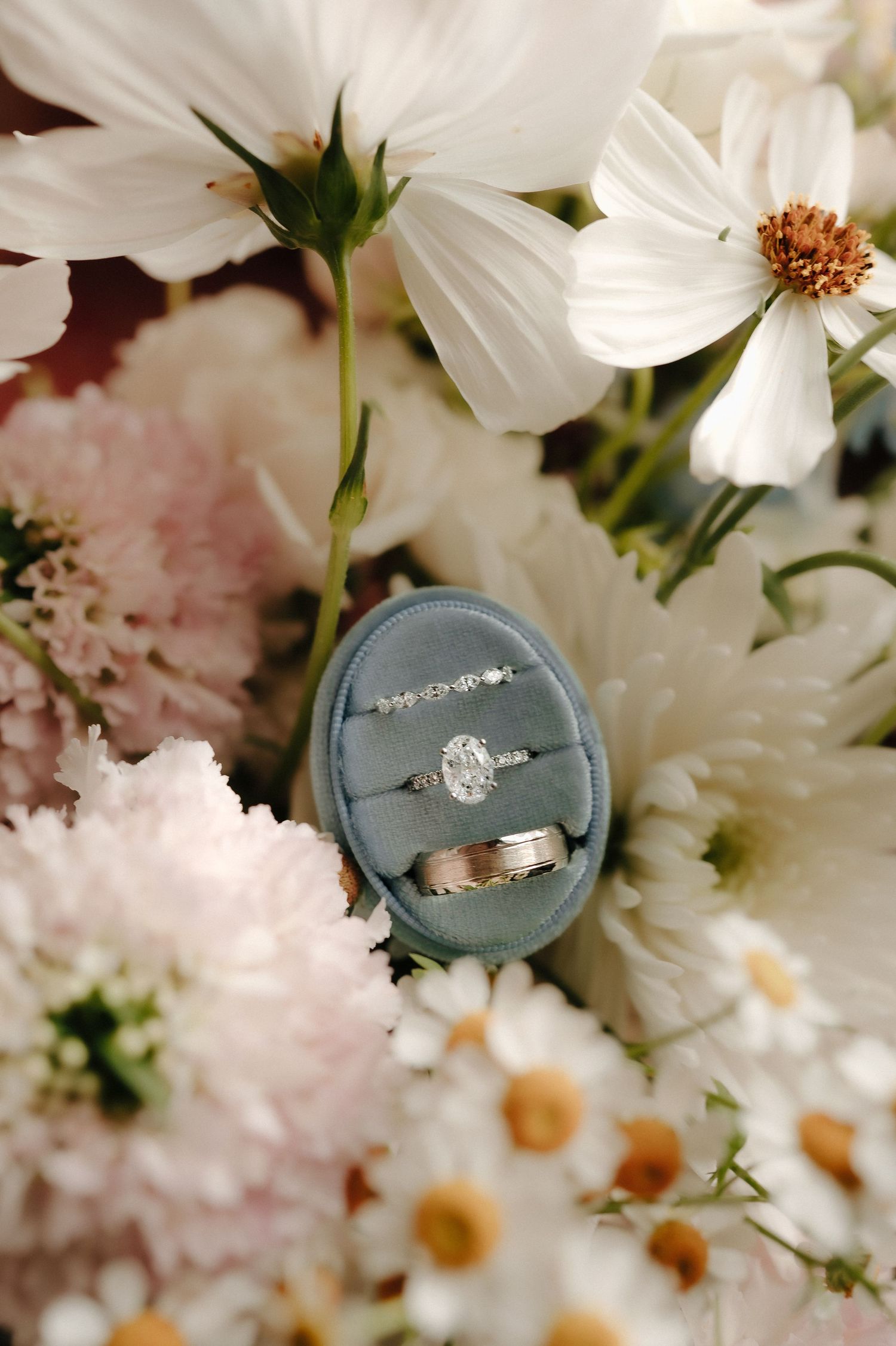 Wedding rings displayed on blue velvet ring box surrounded by white and pink flowers.