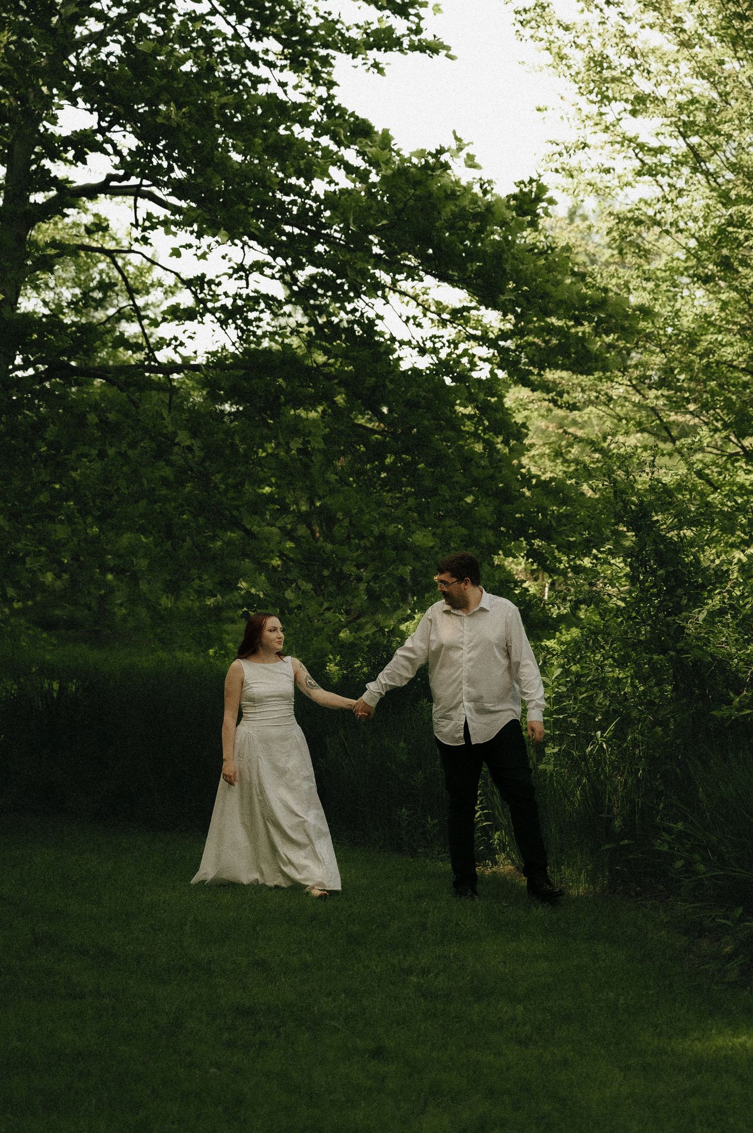 Couple holding hands walking in lush green garden, woman in white wedding dress, man in light shirt and black pants.