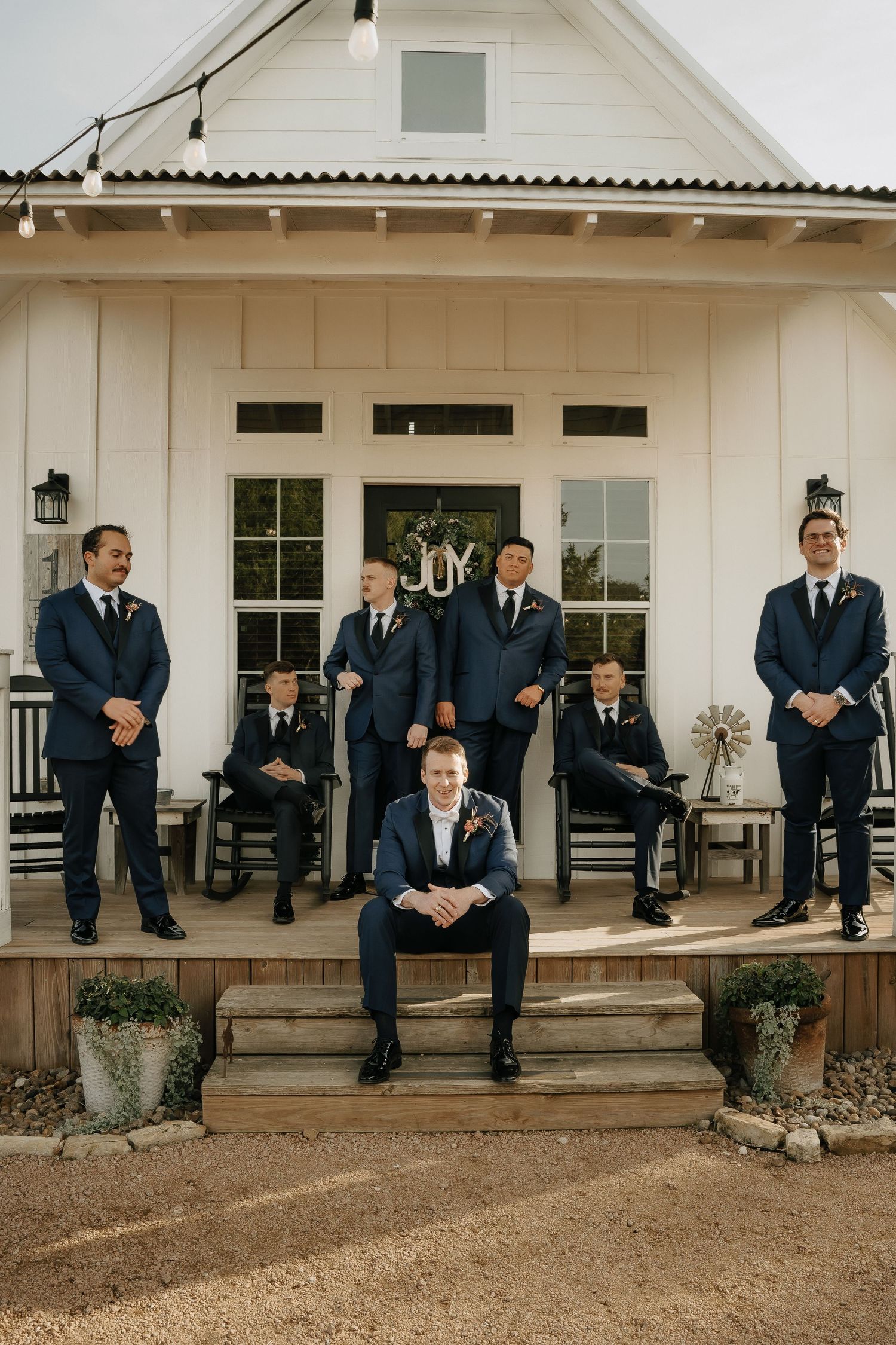 Groomsmen in navy suits sitting casually on white farmhouse porch steps.