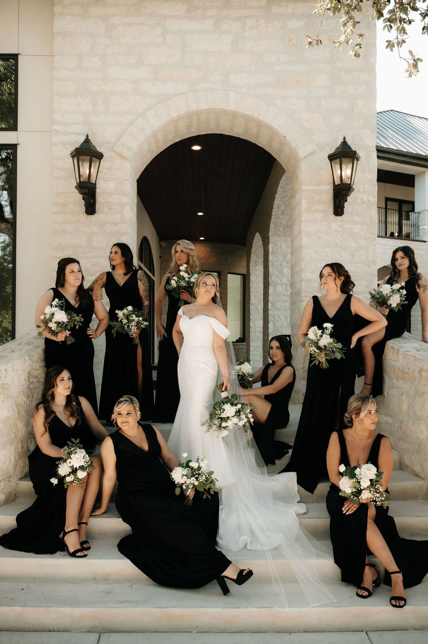 Bridal party in black dresses poses playfully against white stone building.