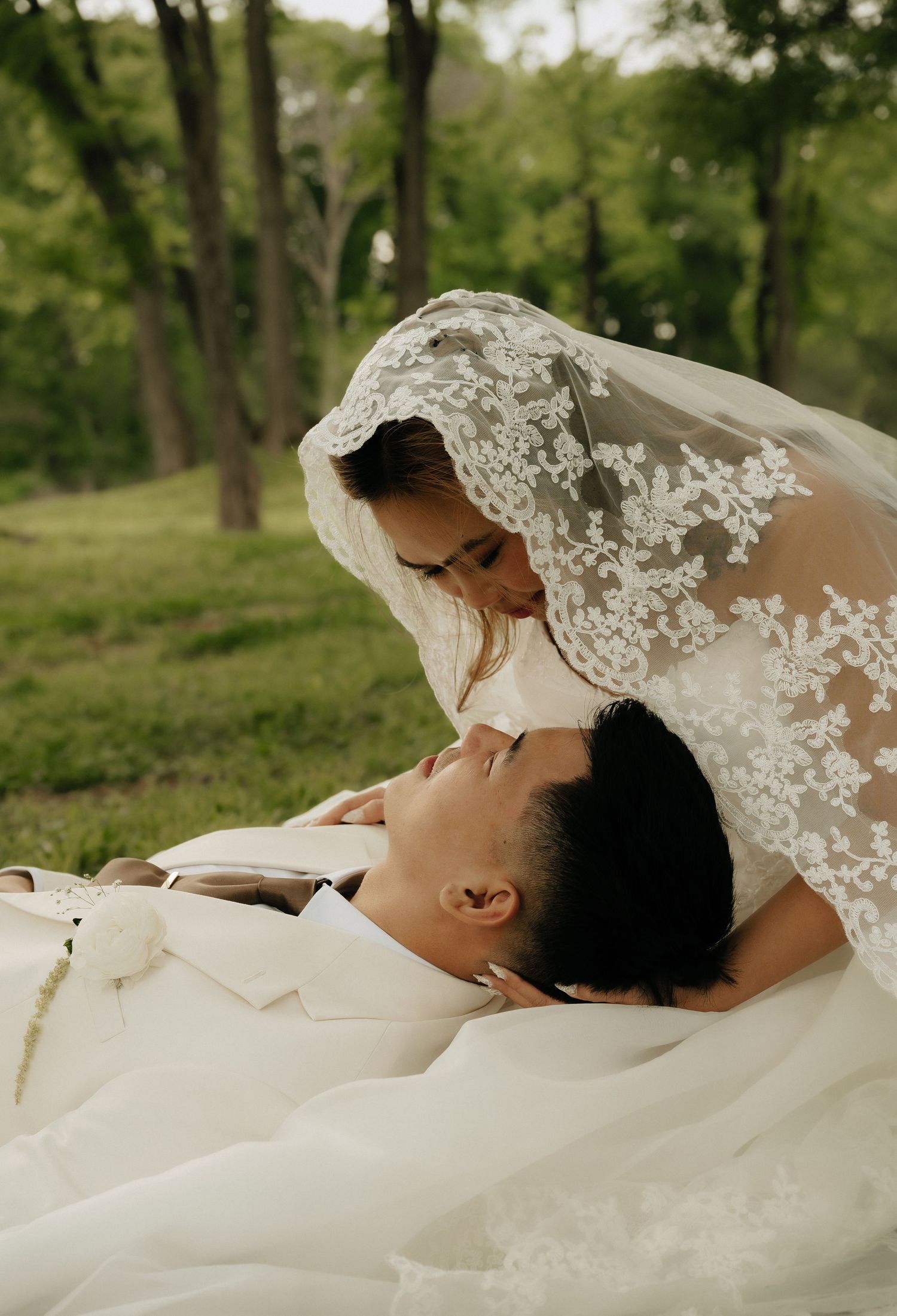 Bride in lace veil leaning over groom lying on grass in romantic outdoor wedding photo.