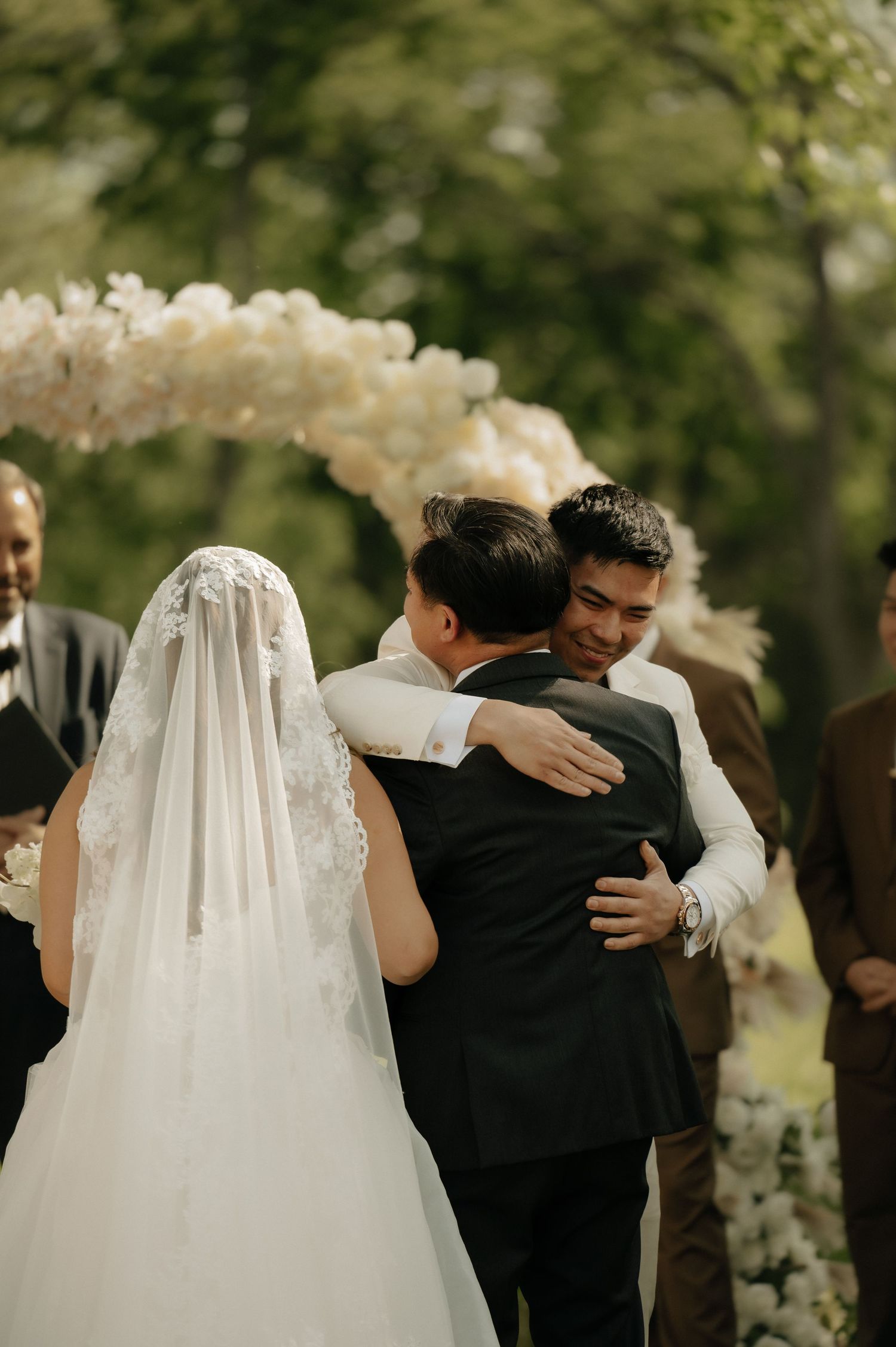 Groom embraces guest at outdoor wedding altar decorated with white floral arch while bride stands nearby.