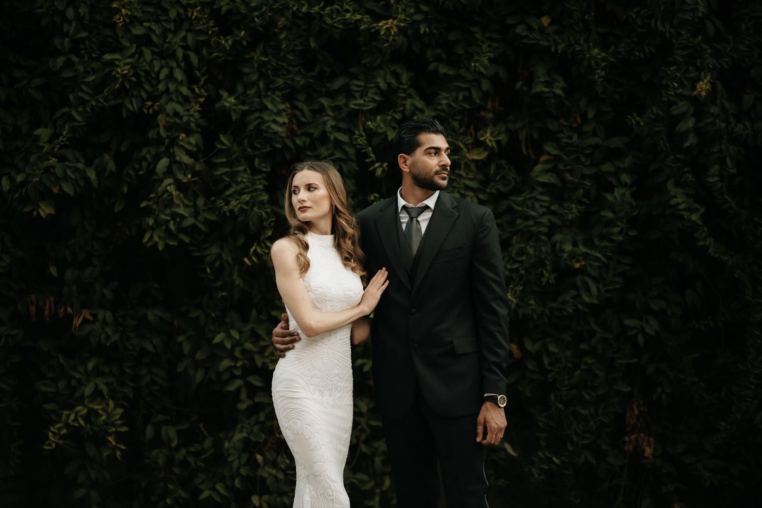 Stylish couple in formal wear poses against lush green hedge backdrop.