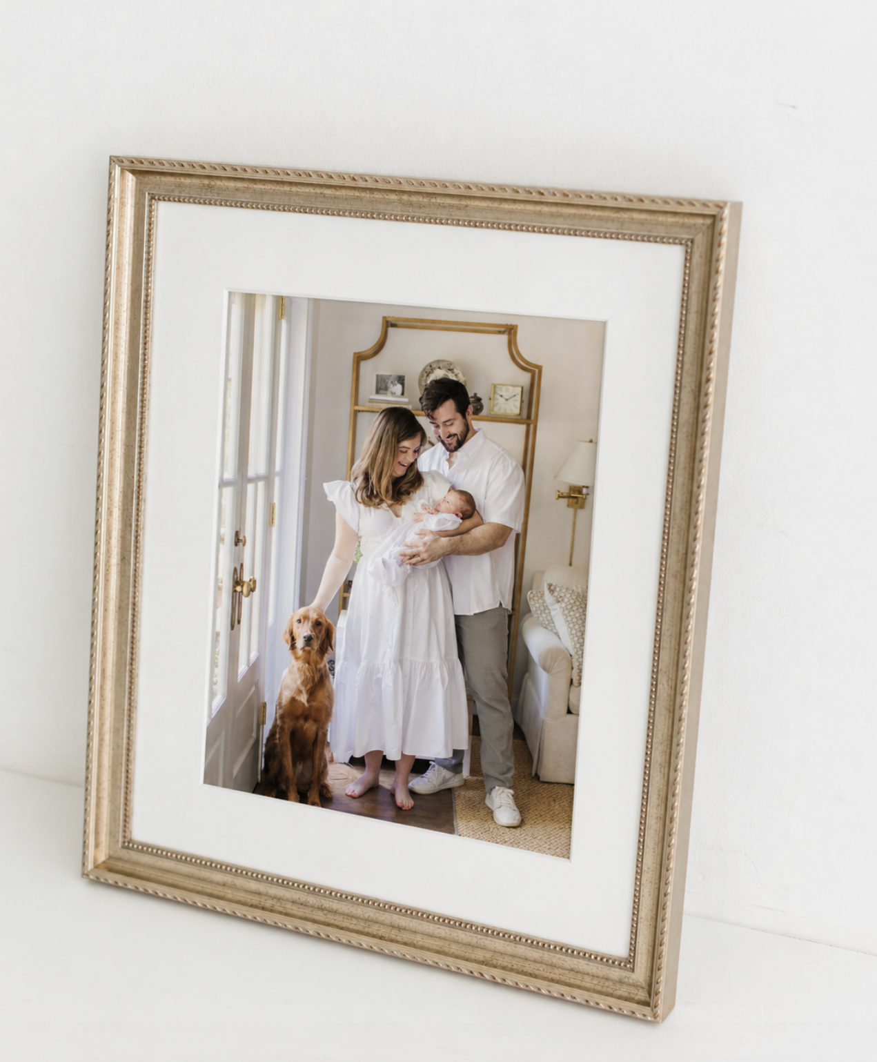 Gold ornate framed family portrait of couple holding newborn baby with golden retriever dog in elegant white room.