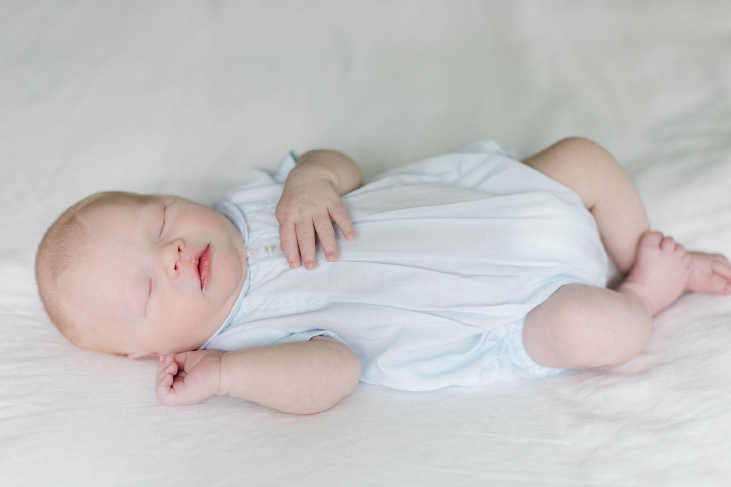 Baby dressed in white lies peacefully on soft white bedding while sleeping.