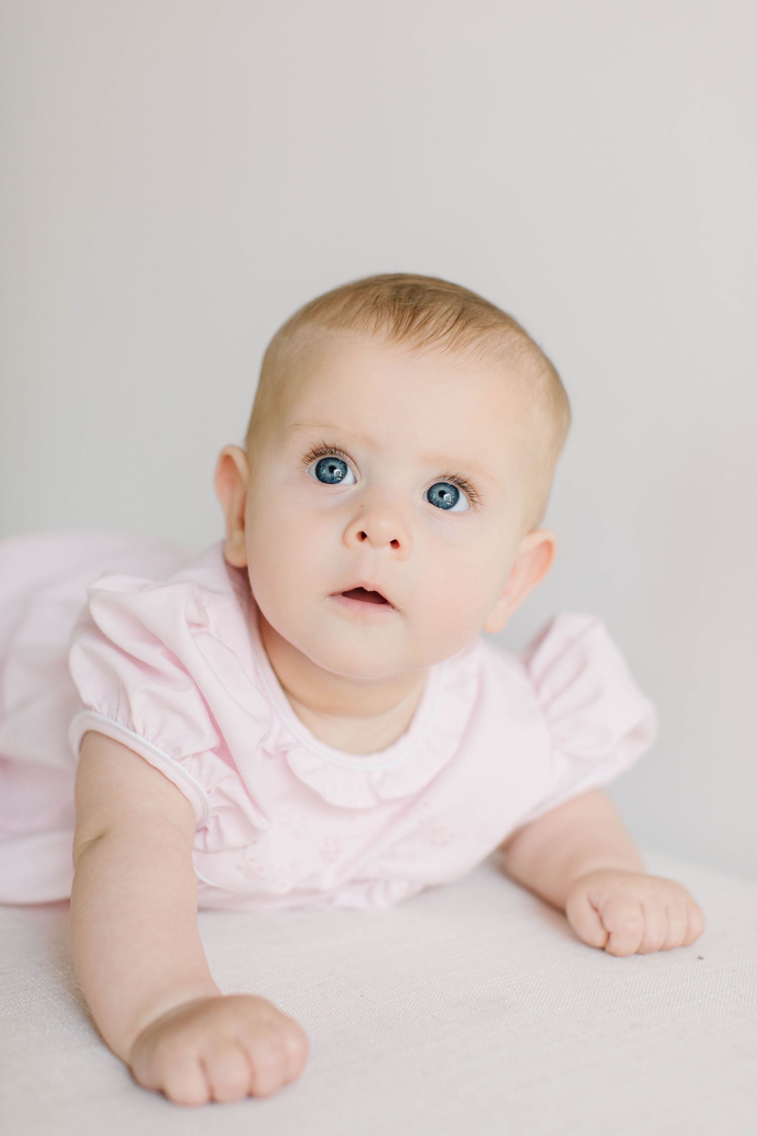 Sweet baby wearing soft pink dress lies on white background showing bright blue eyes.
