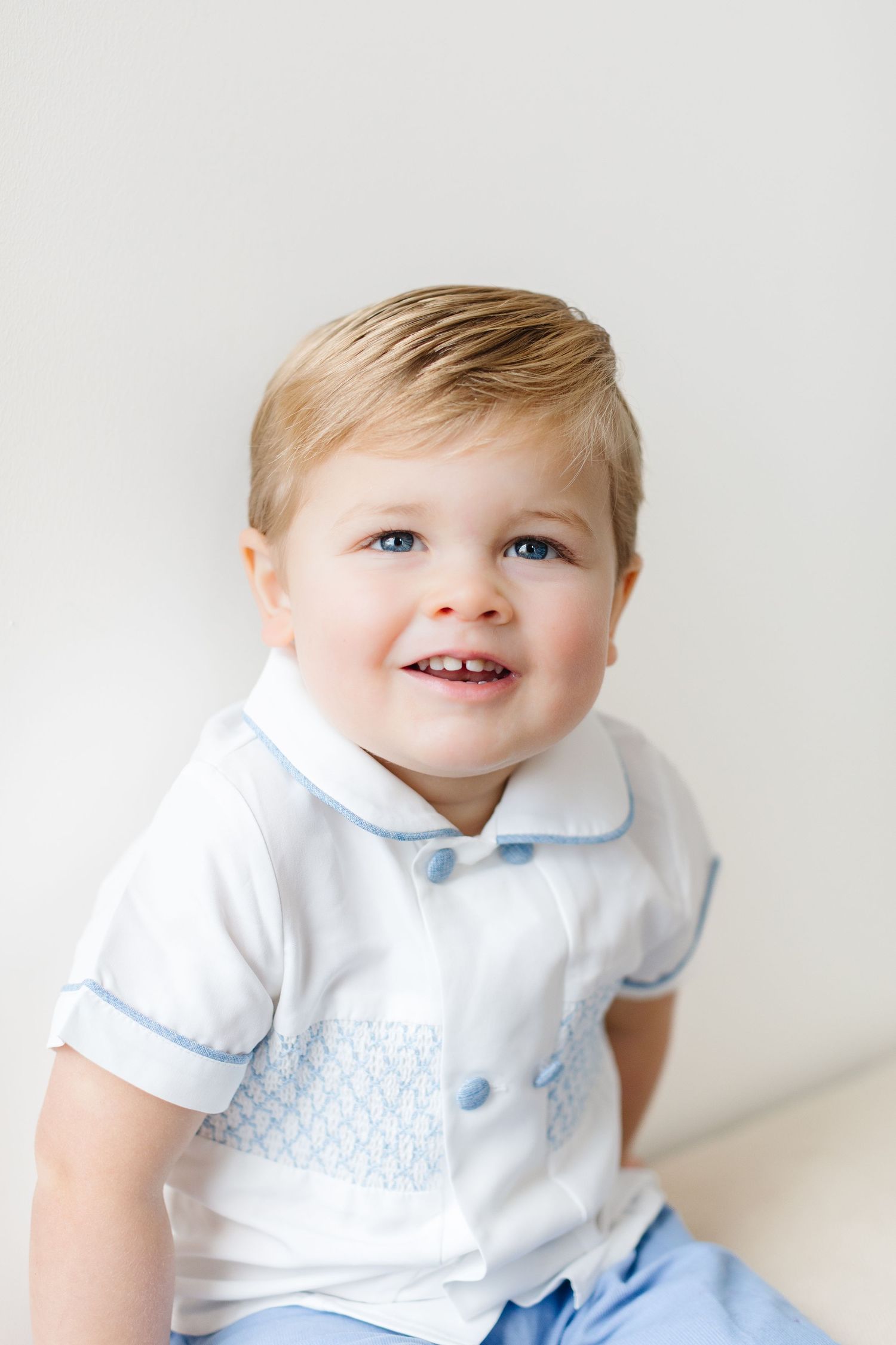 Sweet baby in white collared shirt and light blue bowtie smiles while looking to the side against white background.